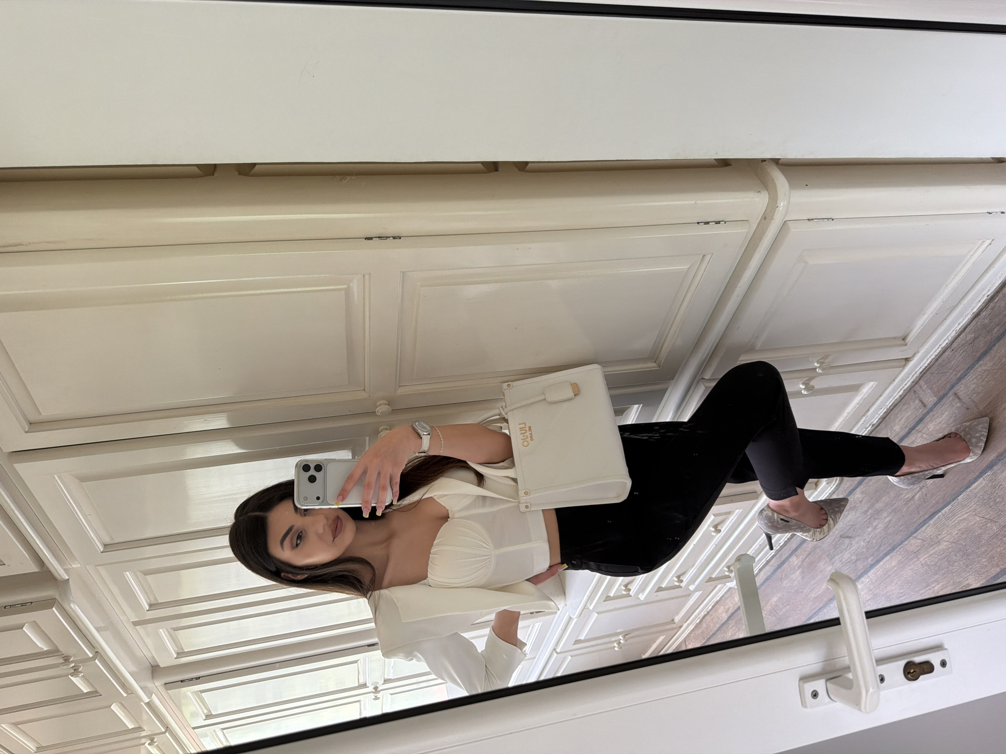 A woman with long, dark hair is taking a selfie in a mirror. She is wearing a white top with a sweetheart neckline and black pants. She is holding a white purse in one hand and a cell phone in the other. She is wearing a watch on her wrist and silver heels. The mirror is reflecting a white wall with cabinets and a wood floor. The location cannot be determined from the image.