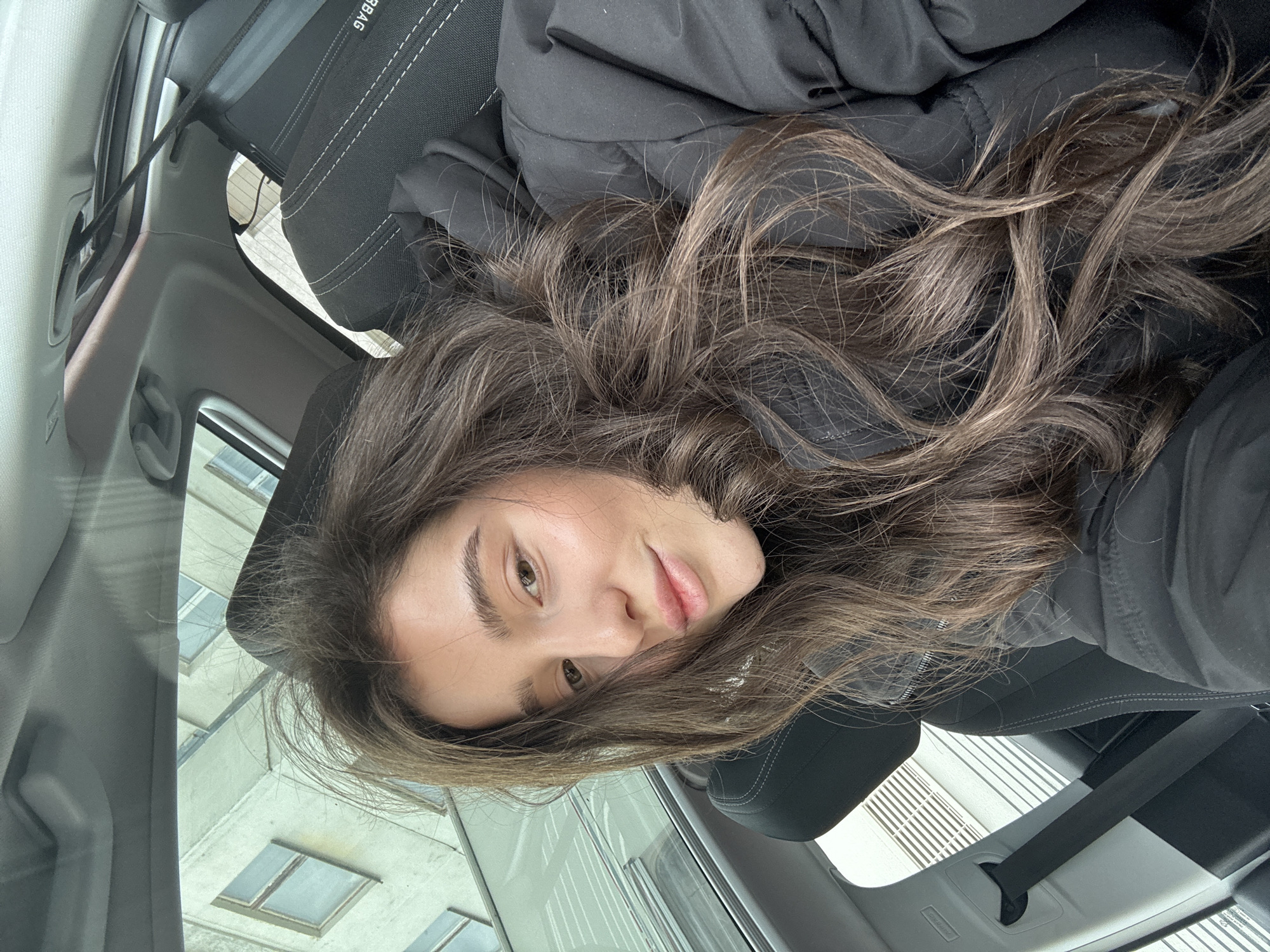 A young woman with long, wavy, brown hair is lying in the passenger seat of a car. She is wearing a dark-colored jacket. Her face is visible, showing light skin, brown eyes, and a neutral expression. The car's interior is visible, including the headrest and seat fabric. Outside the car window, a building with multiple windows can be seen. The location appears to be an urban environment, but specific details about the city are not discernible from the image.