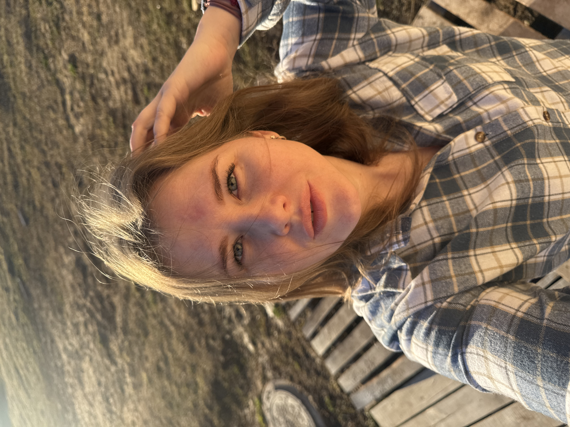 A young woman with light brown hair and blue eyes is lying down outdoors. She is wearing a blue and white plaid shirt. Her right hand is touching her hair. The background appears to be a wooden surface, possibly a deck or walkway, with some grass or dirt visible. The location appears to be near Gornyi (Dvurechenskoe S/P), Russia.