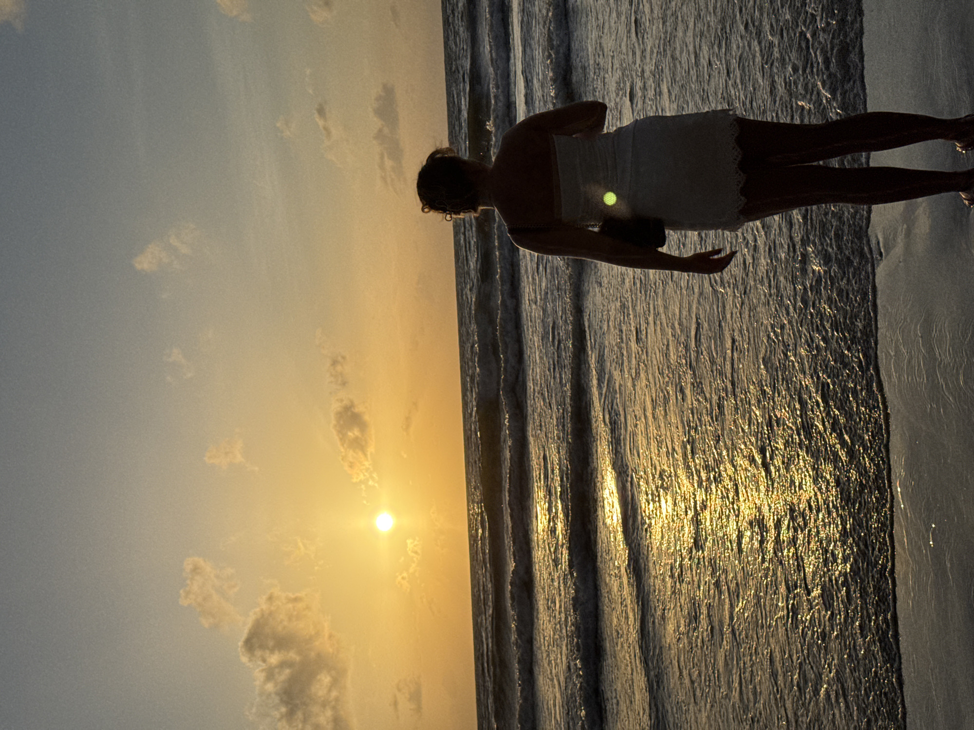 A lone woman stands on a sandy beach in Patong, Thailand, facing the ocean as the sun sets. The sky is a warm gradient of orange and blue, with scattered clouds. The sun, a bright orb, is low on the horizon, casting a golden reflection on the rippling waves. The woman, viewed from behind, wears a white dress and has her hair tied up. She carries a dark bag over her shoulder. The water is a deep blue, with white foam cresting on the waves as they approach the shore. The sand is damp near the water's edge.