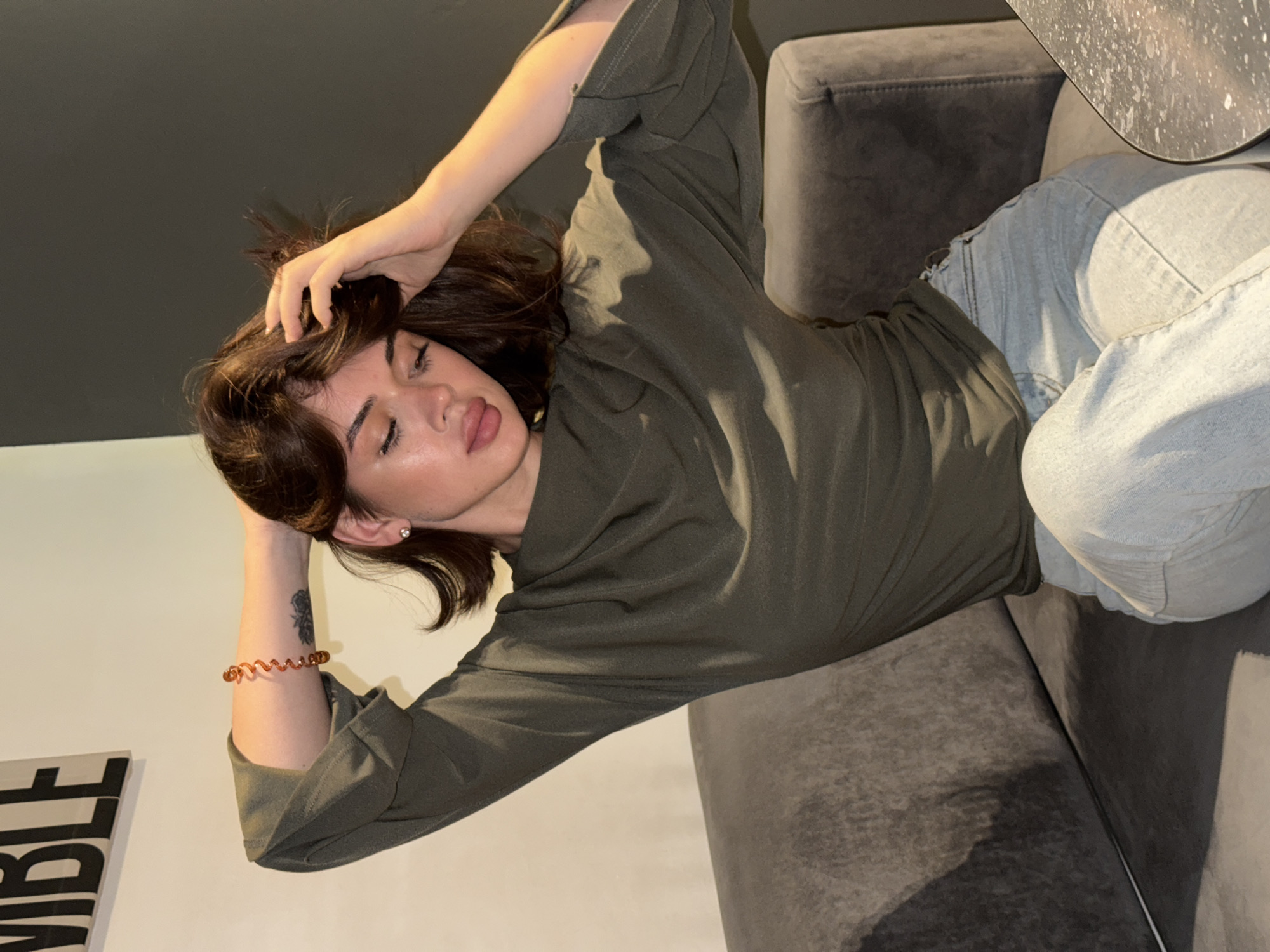 A young woman reclines on a gray upholstered couch against a neutral-toned wall in what appears to be an interior setting. She wears a long-sleeved olive green shirt and light blue jeans. Her left arm is extended above her head, hand touching her hair, while her right arm rests at her side. On her right wrist, she wears an orange bracelet. A dark gray or black ceiling spans the upper left portion of the frame, and a gray table top is visible at the upper right. A partial graphic featuring the word "IBLE" is visible in the lower left corner. The location is presumed to be Chaikovsky, Russia.