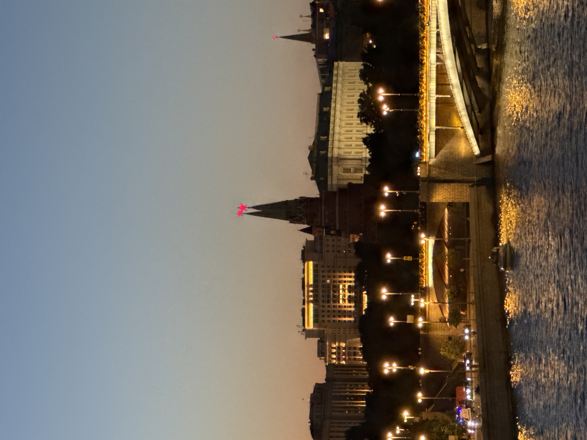 This is an evening photograph of the Moscow Kremlin and the Moskva River. The sky is a gradient of deep blue to pale orange, indicating dusk. The Kremlin's towers, including the Spasskaya Tower, are illuminated, topped with red stars. Several large buildings with illuminated windows are visible within the Kremlin walls. A modern bridge, illuminated with warm lights, spans the Moskva River in the foreground. The river reflects the city lights, creating a shimmering effect on the water. Several streetlights line the embankment and the bridge. A few vehicles, possibly a police car and a truck, are visible on the embankment. A small boat is on the river. The overall scene depicts a well-lit urban landscape at twilight.