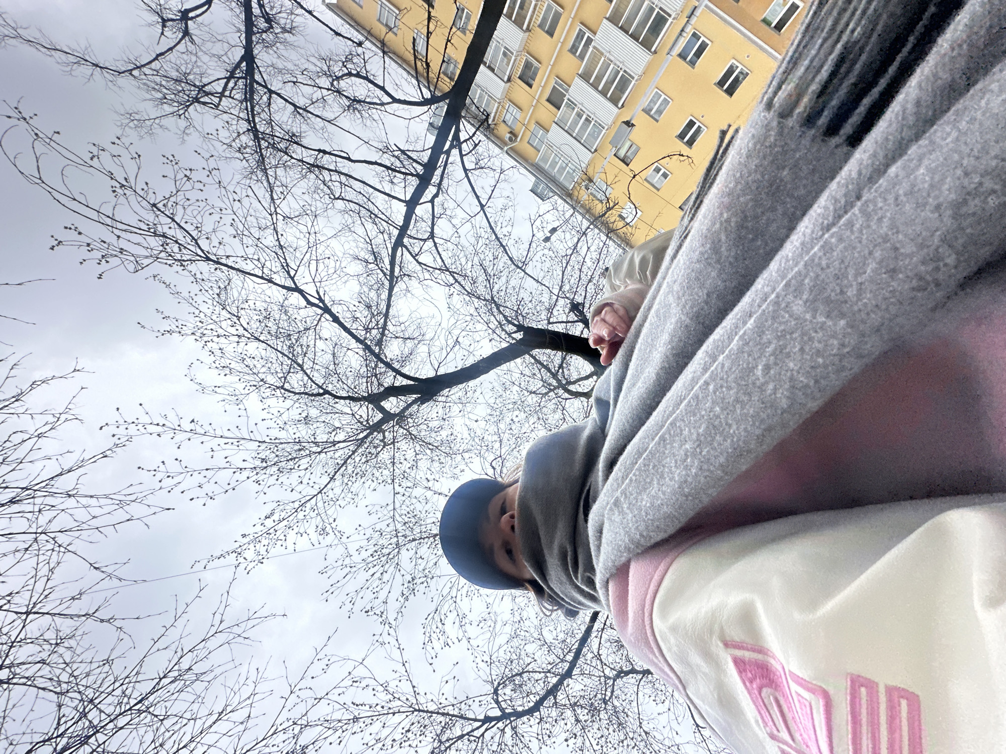 The image is a low-angle, tilted photograph taken from below, looking upwards towards the sky, trees, and a building. The perspective is unusual, with the horizon line sharply diagonal, suggesting a selfie or a shot taken while lying down or holding the camera at an extreme angle.
In the bottom right foreground, a person is partially visible, looking towards the camera. They are wearing a dark blue baseball cap, a grey scarf or blanket wrapped around their neck and shoulders, and a white and light pink top or jacket. On the white section of the top, stylized pink block letters are visible, appearing to be part of a word or logo, possibly "NN" or "MM" or "NY". The person's face is mostly obscured by the cap and the angle, showing only their eyes and nose.
The dominant features of the scene are numerous bare tree branches silhouetted against a light grey, overcast sky. Many of the branches show small buds, indicating late winter or early spring. In the upper background, a section of a multi-story yellow apartment building with numerous white-framed windows and balconies is visible. The building's architecture appears typical of residential blocks found in cities.
The weather is cloudy with diffuse light. The overall mood is calm and perhaps a bit cool, consistent with early spring. Given the provided context, the location is Perm, Russia.