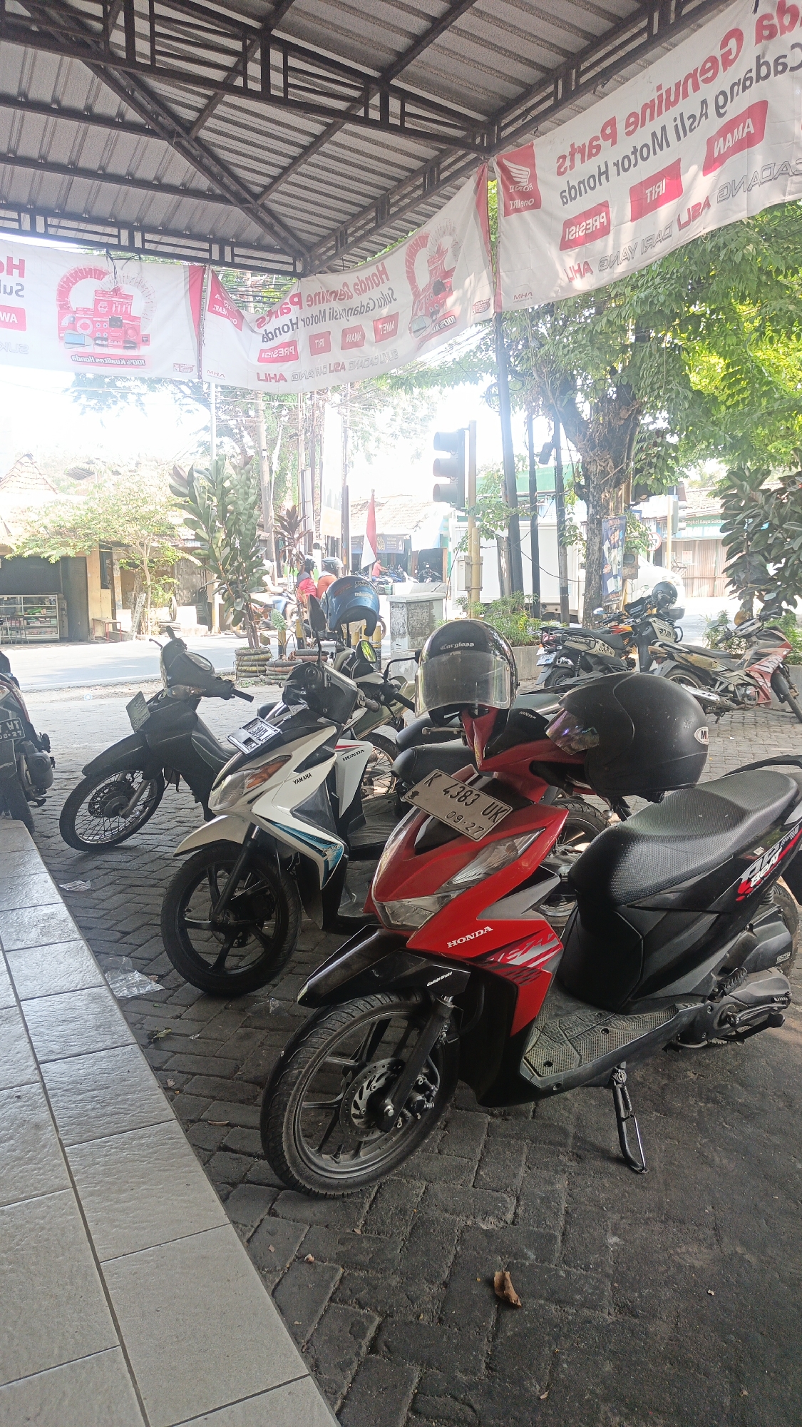 The image depicts a street scene in Mlati Kidul, Indonesia, featuring several parked motorcycles under a covered area. A banner displaying "HONDA GENUINE PARTS" in a mix of English and Indonesian hangs above the motorcycles. The banner also features the Honda logo and the text "HONDA MOTOR ILS A PRODUSEN" and "YAMAHA".

In the foreground, a red Honda motorcycle with a license plate "K 4383 UK" is prominently displayed. Two black helmets rest on the handlebars of this and another nearby motorcycle. To the left, a white Yamaha motorcycle is parked. Further back, more motorcycles are visible, including a black one to the far left and several others in the background.

The ground is paved with interlocking bricks. Trees and buildings are visible in the background, along with a traffic light, suggesting an urban or semi-urban environment. The lighting indicates it is daytime, with sunlight filtering through the trees and the covered area. No people are clearly visible or engaged in any action. The overall scene is static, focusing on the parked vehicles and the advertising banners.