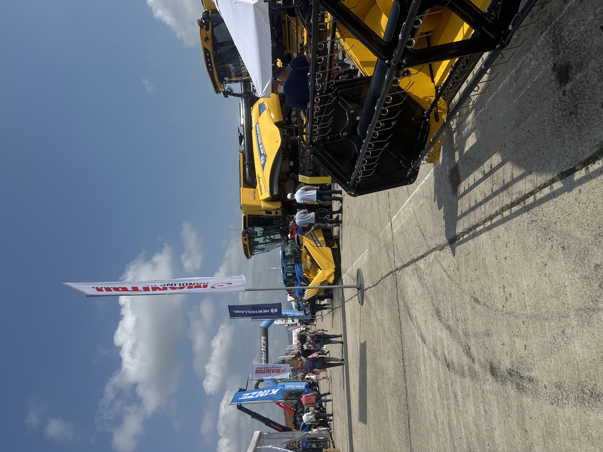 This media file depicts an outdoor agricultural machinery exhibition or trade show, likely in Stara Zagora, Bulgaria, under a clear, bright blue sky with scattered clouds. The scene is set on a wide paved area where various large agricultural machines, primarily in yellow and blue, are displayed.
In the foreground, a prominent yellow combine harvester with a complex black cutting mechanism is visible, alongside several individuals observing it. Further down the exhibition aisle, more machinery, including tractors and what appears to be a telehandler, are on display. Numerous people, mostly adults dressed casually, are seen walking around the exhibits, indicating an active event.
Several banners and flags line the area, featuring brand names and event titles. Visible text includes "NEW HOLLAND," "MANITOU" (with the slogan "HANDLING YOUR WORLD"), "KINZE," and "InterAGRI," which also appears on a blue inflatable archway. A specific detail on one yellow machine reads "RÖTOR," and a faint Cyrillic text "РОДА П" is also discernible. The bright sunlight casts distinct shadows on the ground, suggesting a daytime event. The overall atmosphere appears engaging and busy, characteristic of a trade fair.