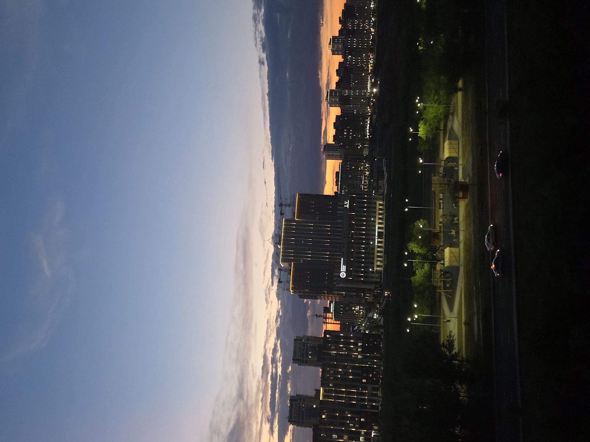 The image captures a twilight scene in the city of Karaganda, Kazakhstan. A modern cityscape with numerous illuminated buildings is visible against a dusky sky painted with hues of blue, orange, and gray clouds. The buildings, many of which appear to be residential complexes or office towers, are lit up with a mixture of white and orange lights. Construction cranes are present on some of the taller structures, suggesting ongoing development. In the middle ground, a park area is illuminated by streetlights, with several structures that resemble playground equipment or small buildings. A road runs in the foreground, where two cars are visible. The overall atmosphere is one of a developing urban environment transitioning from day to night.