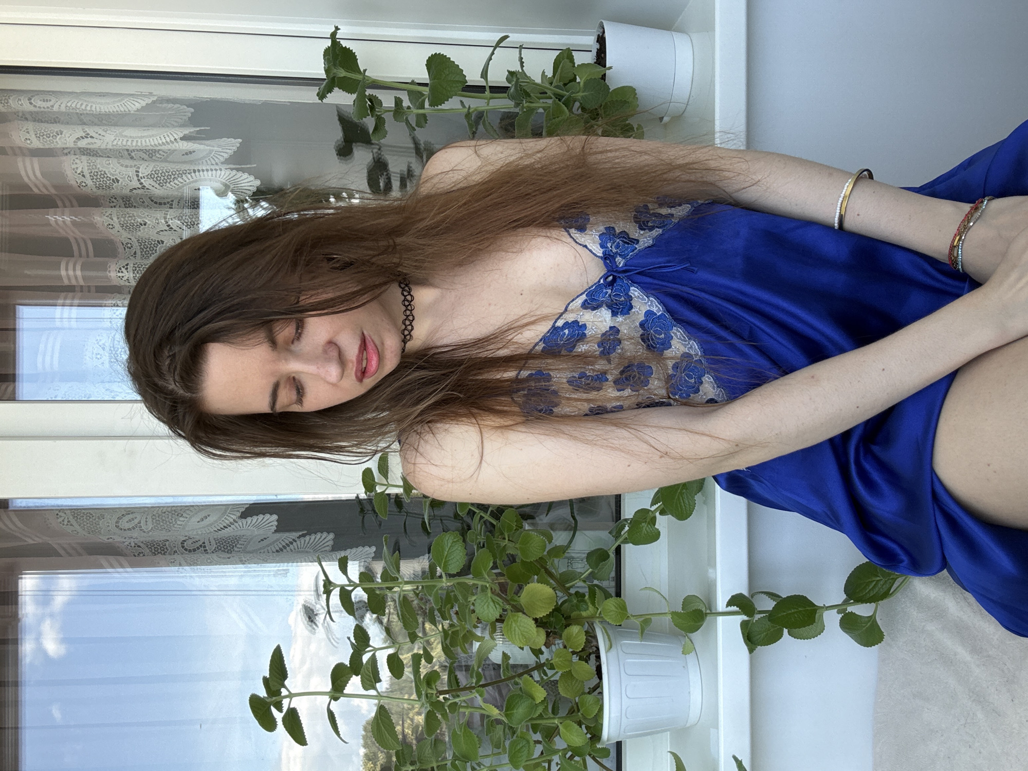 A young woman with long brown hair, wearing a blue satin slip with floral lace detailing, is seated indoors near a window. She is looking down with her eyes closed. Green plants in white pots are visible on the windowsill, and the window reflects a bright sky with clouds. The woman is wearing a black choker necklace and bracelets on her left wrist. The background consists of the window, lace curtains, and a wall. The location is described as Unknown, Unknown.
