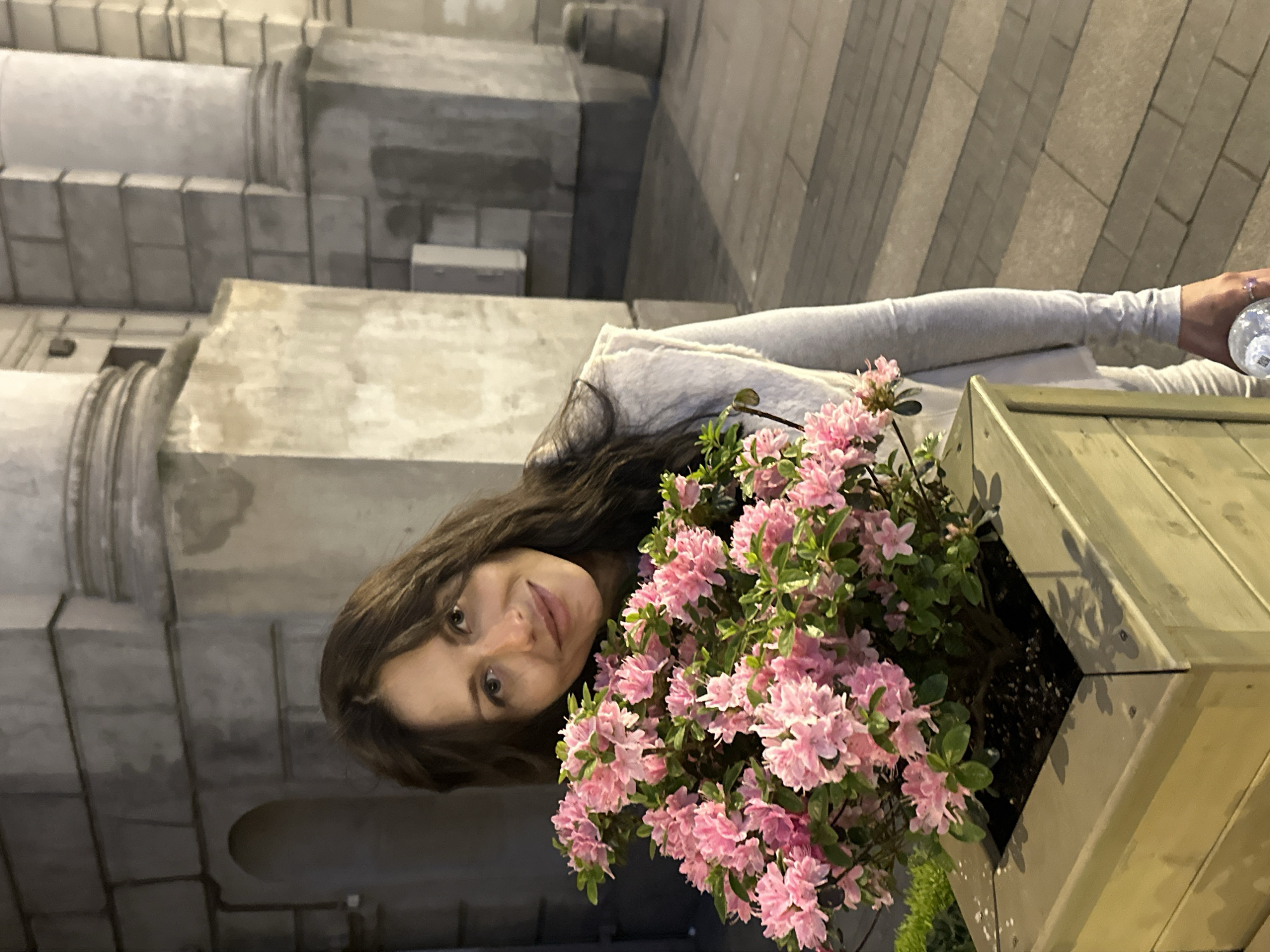 The image, originally rotated, captures a young woman posing alongside a vibrant planter of pink flowers in an urban setting during the evening or night. The woman, with long, dark, wavy hair, smiles gently as she looks directly at the camera. She is wearing a light-colored top or jacket, and her left hand, adorned with a ring, holds a clear plastic water bottle.
Adjacent to her is a large wooden planter box, constructed with a distinct diagonal pattern, filled with numerous clusters of bright pink blossoms, likely azaleas, which appear well-lit against the dim backdrop.
The background features impressive stone or concrete architecture, characterized by large, light-toned blocks and decorative elements resembling columns or pilasters, indicative of a grand, historic building. The ground is paved with rectangular blocks. The scene's overall lighting suggests artificial illumination in a low-light environment, creating a serene atmosphere typical of a European city street at night. This architectural style aligns with the provided context of Saint Petersburg, Russia. No text is visible in the image.
