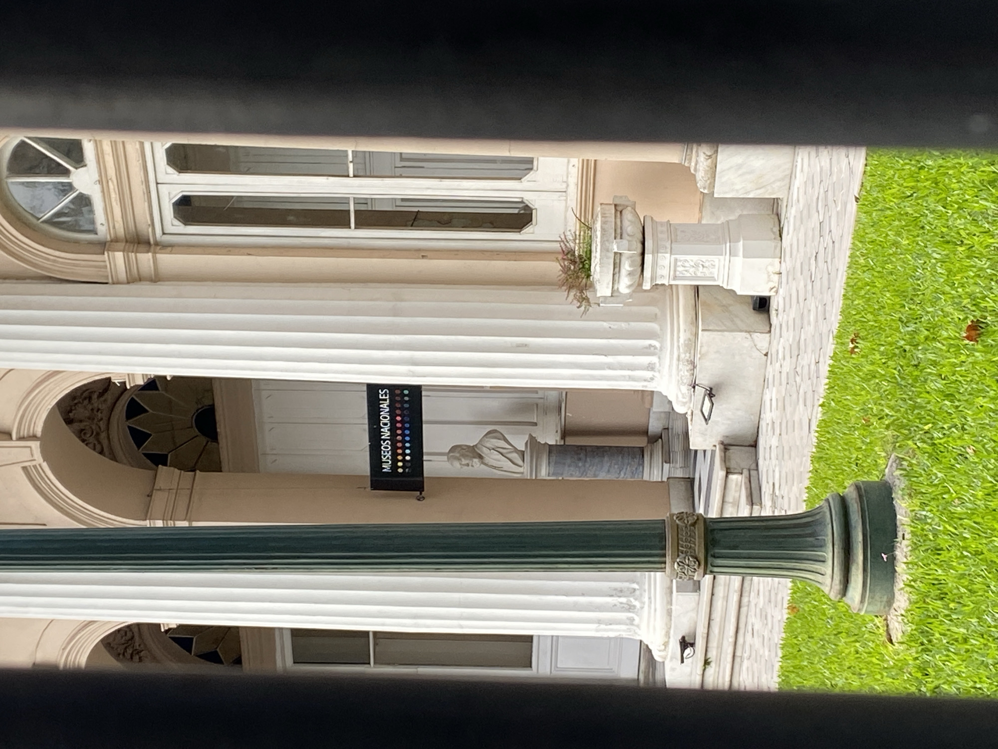 The image shows the façade of the Museo Nacionales in Buenos Aires, Argentina. Visible are classical architectural elements including white columns, arched windows, and stone detailing. A vertical banner with the words "Museos Nacionales" is displayed. A sculpture of a person is visible in the background. The exterior includes a strip of green grass. The photograph appears to have been taken through a partial barrier, obscuring the top and bottom of the frame.