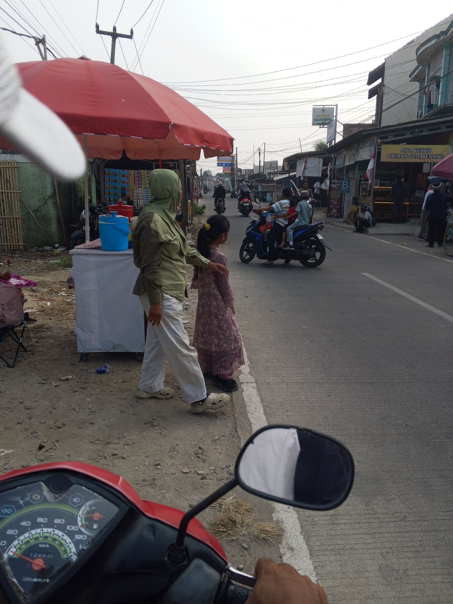 The image depicts a street scene in Ciruas, Indonesia, with several people and vehicles present. A woman wearing a green hijab and a patterned olive shirt stands next to a young girl in a pink patterned dress. They are positioned on the side of a road, near a white cart with a blue cooler on top, covered by a large red umbrella. The street is paved and has white lane markings. On the road, a blue motorcycle carries two people, a man and a child wearing a cap. Several other motorcycles and pedestrians are visible further down the street, along with buildings lining the road, some with signs. The environment suggests a commercial area or a roadside market, with the sky overcast. The foreground shows a red motorcycle's dashboard, indicating the perspective of someone on the vehicle.