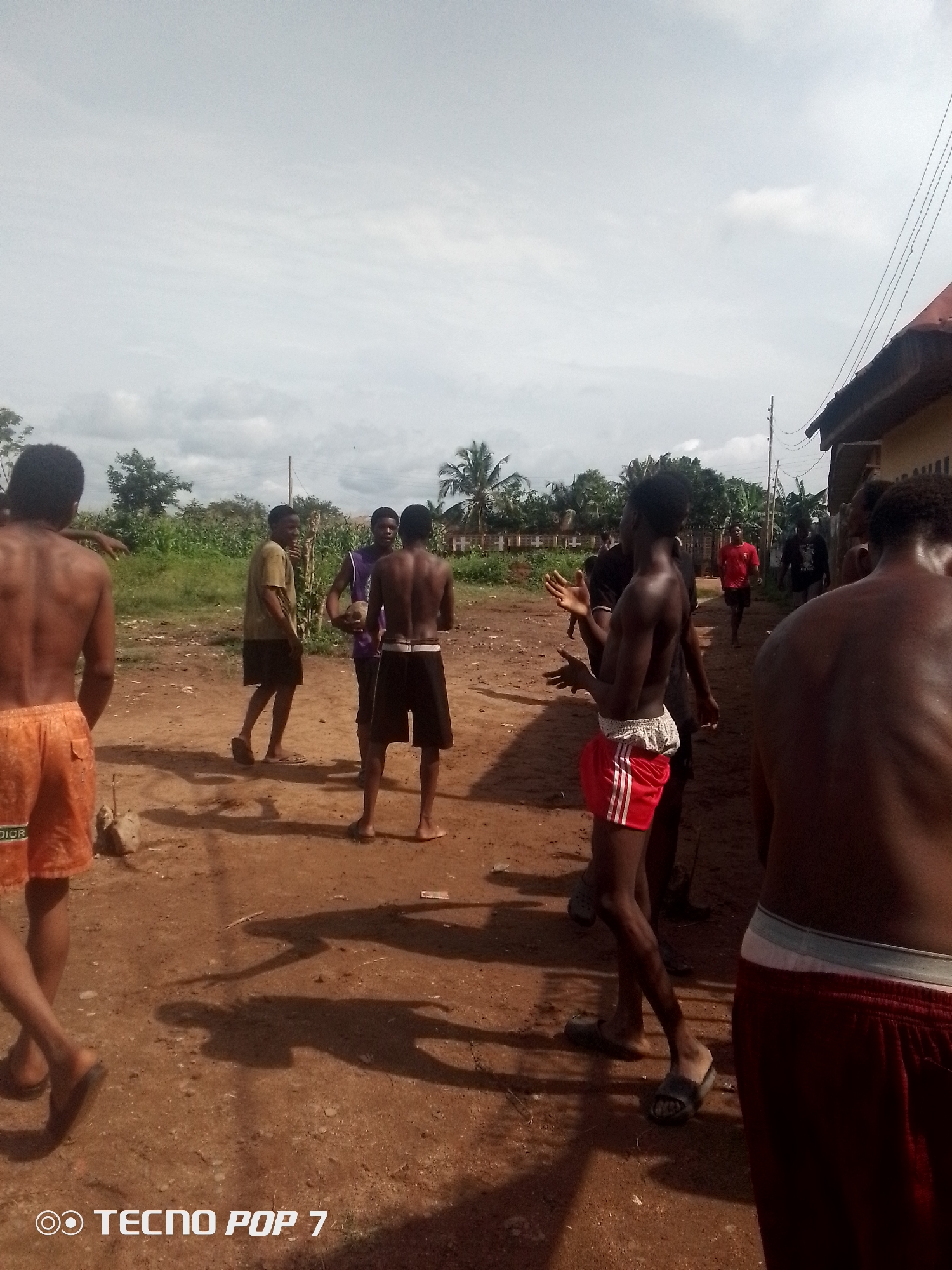 A group of young men are gathered outdoors in a dusty, outdoor area with a building visible on the right. The men are mostly shirtless, wearing shorts. One man in the foreground on the left is wearing orange shorts, while another in the center foreground is wearing red and white striped shorts. There are trees and vegetation in the background, along with utility poles and wires. The ground is dirt and appears dry. The sky is overcast with some clouds. The scene suggests a casual, possibly warm-weather gathering.