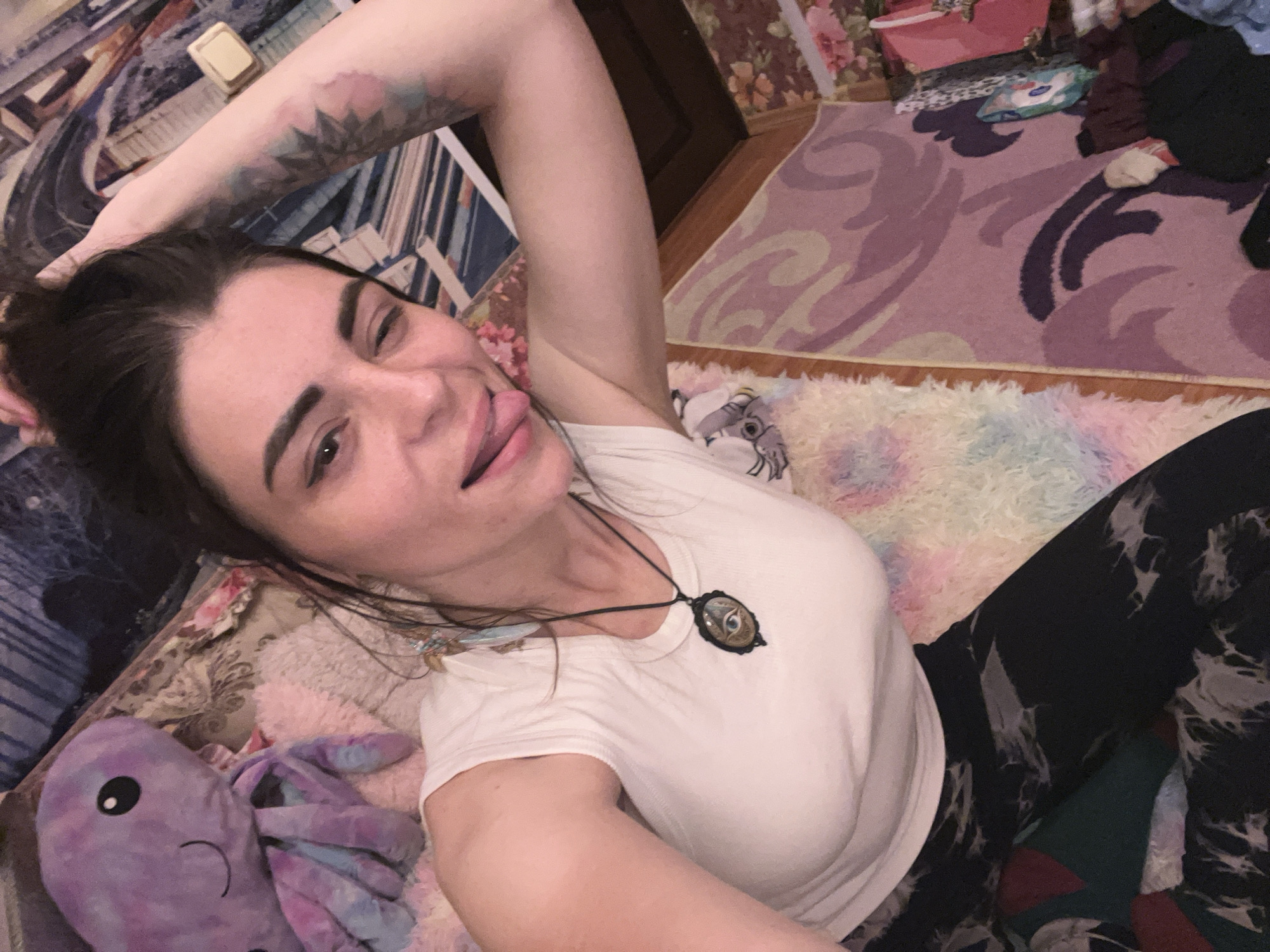 The image shows a woman with dark hair, wearing a white sleeveless shirt and dark patterned leggings. She is lying down, with her right arm raised, holding her hair. She is winking and sticking out her tongue. To her left, there is a large purple stuffed octopus. The background consists of patterned blankets and rugs. There are no discernible features that would indicate the specific location of Krasnoe-na-Volge, Russia.