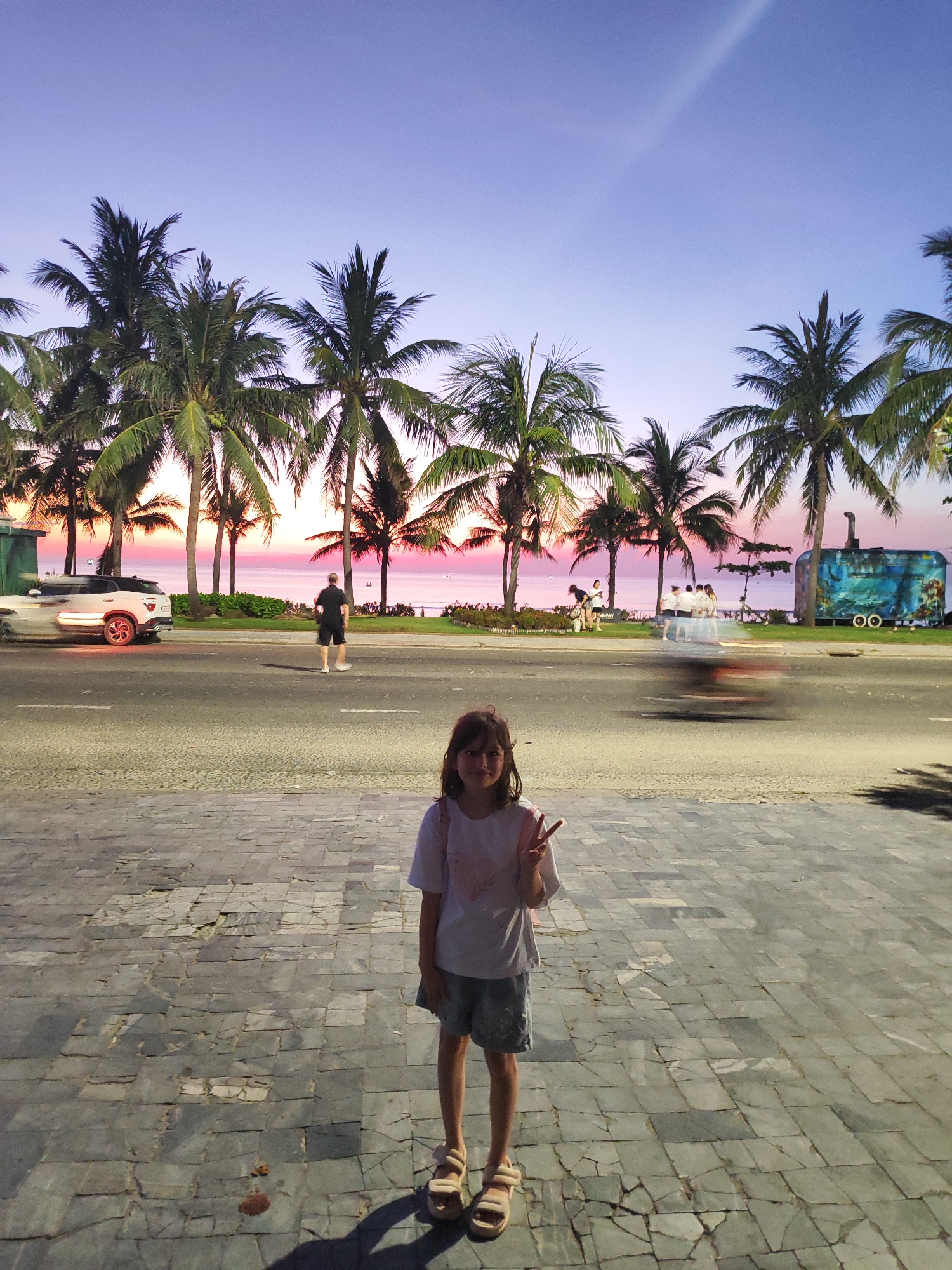 A girl stands on a paved walkway, making a peace sign with her left hand. She wears a short-sleeved white T-shirt, denim shorts, and sandals.
Behind her is a road with moving vehicles and pedestrians. Beyond the road is a line of palm trees against a pink and purple sunset sky. The sea is visible between the palm trees.
A white SUV is on the left side of the road, and a person is walking on the roadside. Further along, a group of people stand near the water, and a food truck is parked on the right.
The location is in Đà Nẵng, Vietnam.