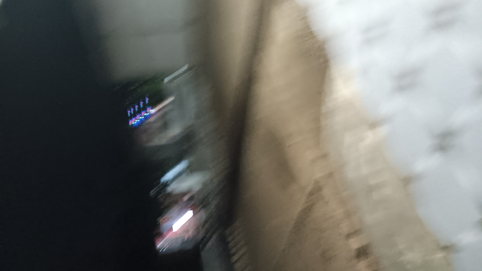 The image is a blurry, low-light photograph taken at night, likely from a moving vehicle. The scene appears to be an outdoor urban environment. In the background, there are streaks of colorful lights, suggesting vehicles or decorative lighting. The ground in the foreground is a light brown, dusty surface, possibly a road or a construction area. There are indistinct shapes and forms throughout the image, making specific identification difficult. The overall impression is one of motion and poor visibility, common in nighttime photography with a moving subject or camera. There is no discernible information to indicate the specific location within Sonobekel, Indonesia.