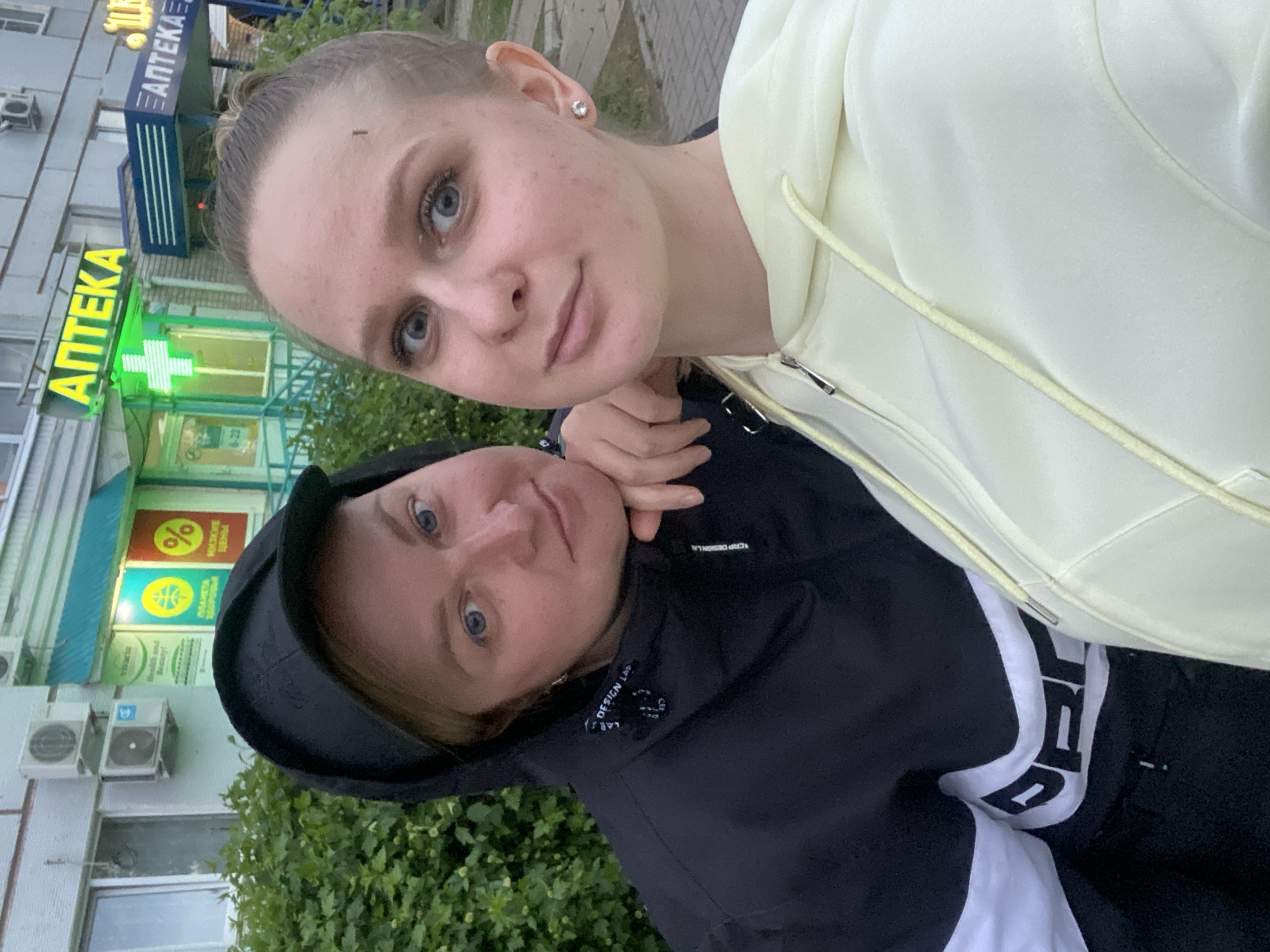 Two women pose closely in a photograph that seems to be taken outdoors. One is standing and wears a light yellow zip-up jacket. The other is wearing a black hooded jacket, with the hood up, and what appears to be white lettering on the front. The background includes a building with a sign that reads "АПТЕКА" (Pharmacy). There is also a green cross visible, which often indicates a pharmacy in Russia. Additional signage includes the words "Vivacia Health and Beauty!", "ПЛАНЕТА ЗДОРОВЬЯ", "НИЗКИЕ ЦЕНЫ", and a percent sign "%". Air conditioning units are visible on the side of the building. Based on the information given, it can be inferred that the location is in Chaikovsky, Russia, near a pharmacy.