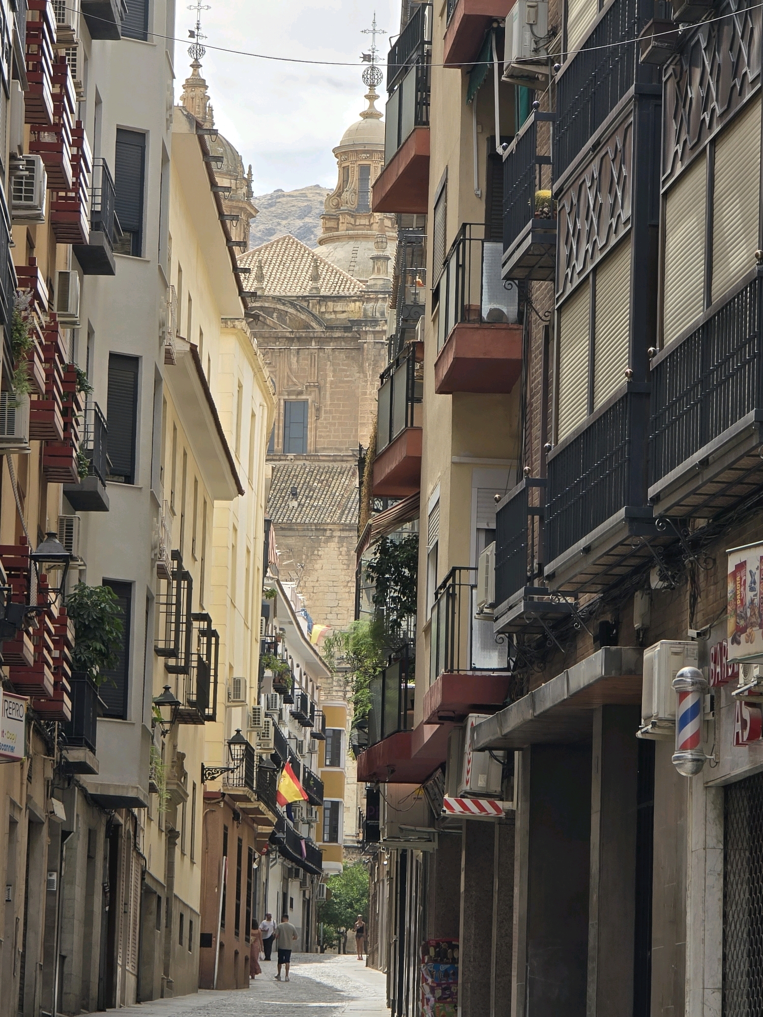 A narrow, cobbled street is lined with tall, multi-story buildings. The buildings are a mix of styles and colors, with some featuring balconies and awnings. Several air conditioning units are visible on the building facades. In the distance, a large building with domes and spires can be seen, possibly a church or cathedral. A Spanish flag is hanging from a balcony on the right side of the street. There are a few people walking in the street, and a barber pole is mounted on the side of a building on the right. The sky is overcast.