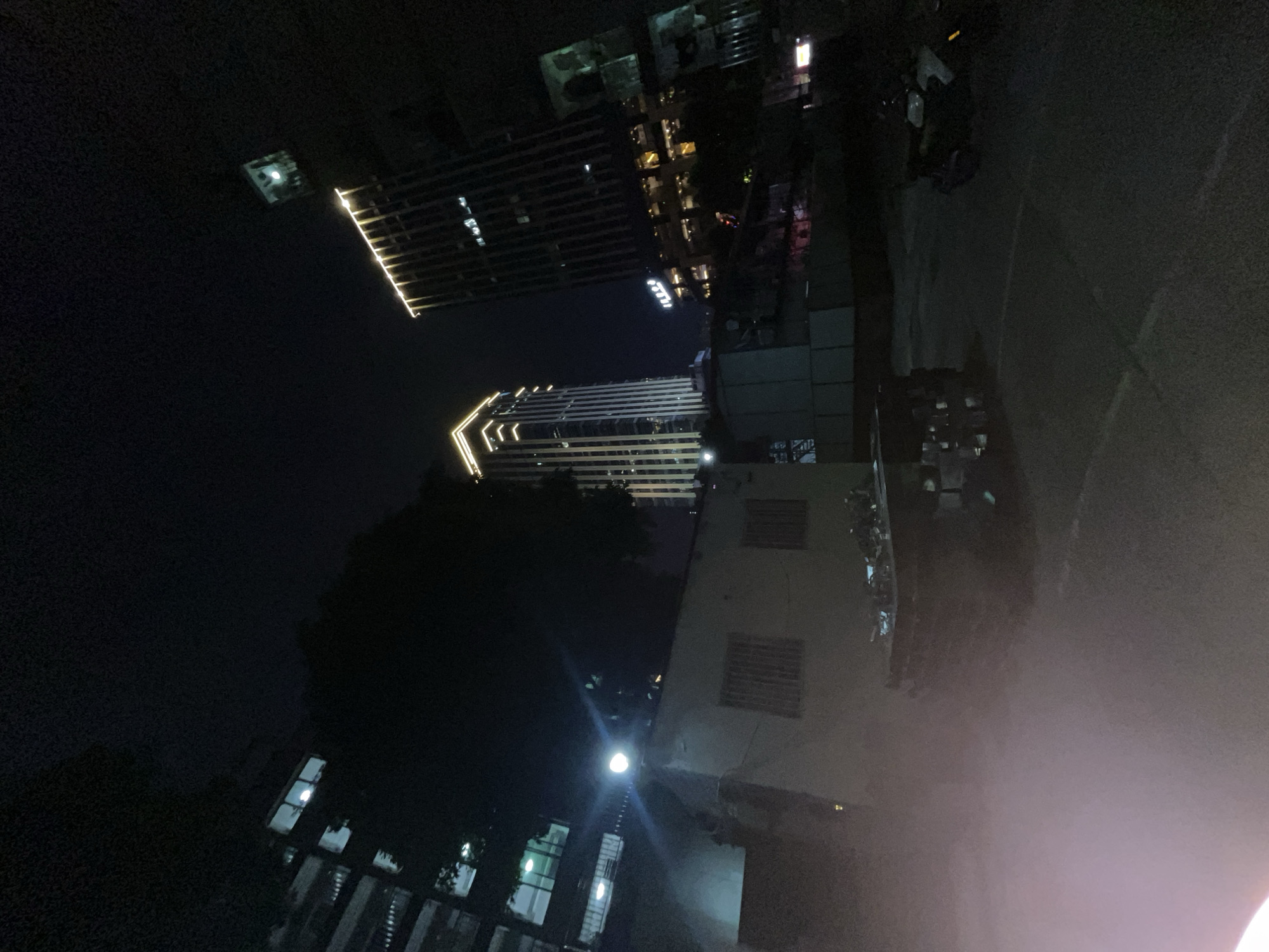 This is a low-light, nighttime photograph taken from a street-level perspective in the Yuzhong District of China. The scene features several buildings, including two prominent skyscrapers with illuminated decorative lighting at their summits. One skyscraper on the left is lighter in color and has a geometric, tiered design at the top, while the skyscraper on the right is darker and features vertical lines. In the foreground, a low-rise building with barred windows is visible, partially obscured by a tree. A bright spotlight shines from the roof of this low-rise building, casting a beam downwards. In the middle ground, there appears to be a collection of stacked objects, possibly crates or furniture, on a table or flat surface. A motorcycle is parked on the right side of the image. The ground is paved, suggesting a street or parking area. A faint haze or mist is present in the lower left portion of the frame. The overall impression is of an urban environment at night, with a mix of residential and commercial structures.