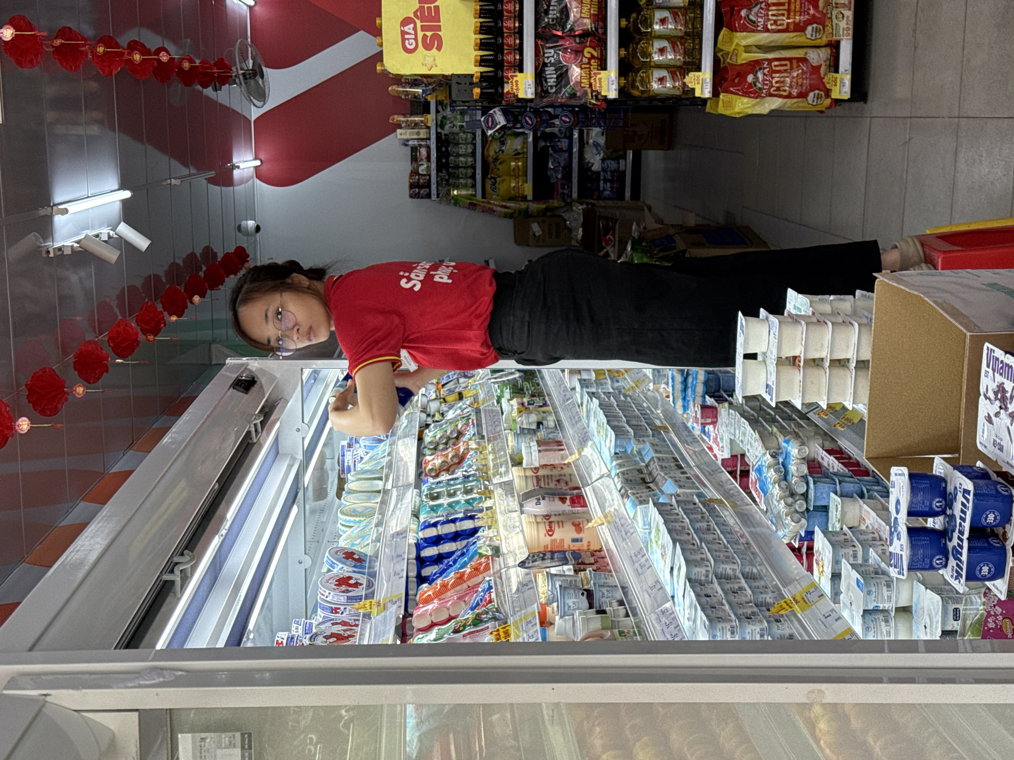 A young Vietnamese woman wearing a red shirt and black pants is standing on a red stool inside a convenience store in Bac Nha Trang Ward, Vietnam. She is looking to her right, towards the camera. The store is filled with shelves of products, including yogurts, canned goods, and bottles of oil. Red lanterns are hanging from the ceiling, suggesting a festive occasion. The woman appears to be a store employee, as she is wearing a uniform and seems to be restocking shelves.