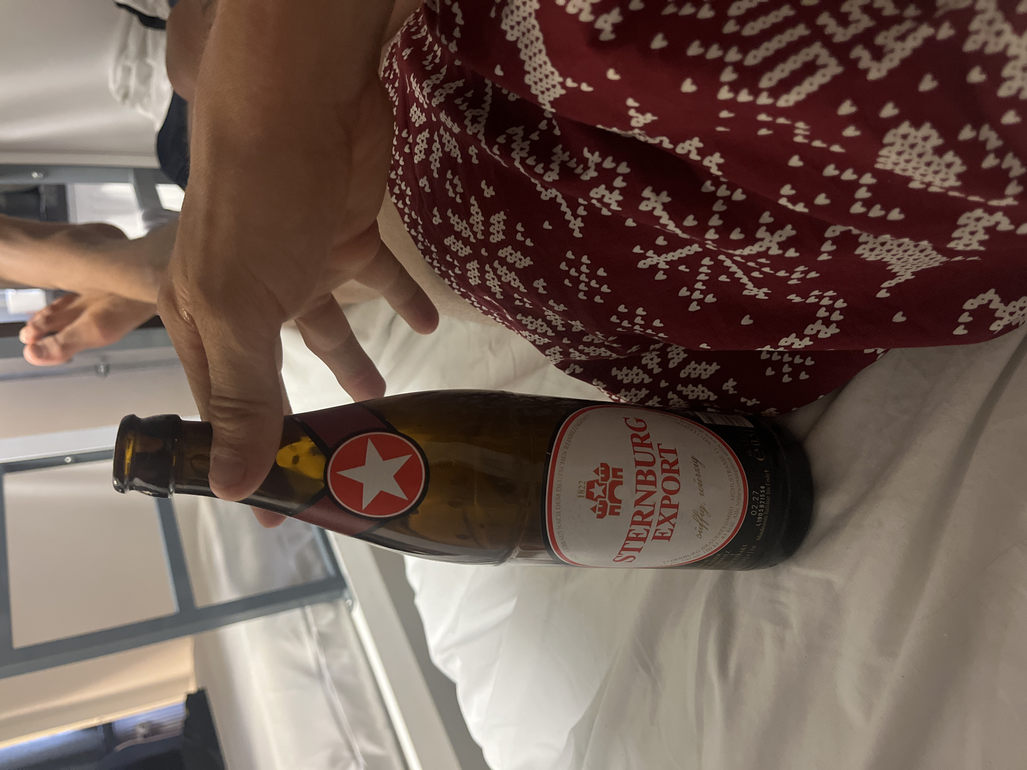 In the image, a person's hand is holding a brown glass bottle of Sternburg Export beer. The bottle has a red and white label with a star and the text "STERNBURG EXPORT". The person is sitting on a bed with white sheets. In the background, there are bunk beds, suggesting the location is likely a hostel or dormitory. The image also shows a person's feet crossed in the background, indicating the presence of at least one other person. The overall scene appears to be indoors, in a casual setting. Without further context or recognizable landmarks, it is impossible to definitively place this image in Berlin, Germany.