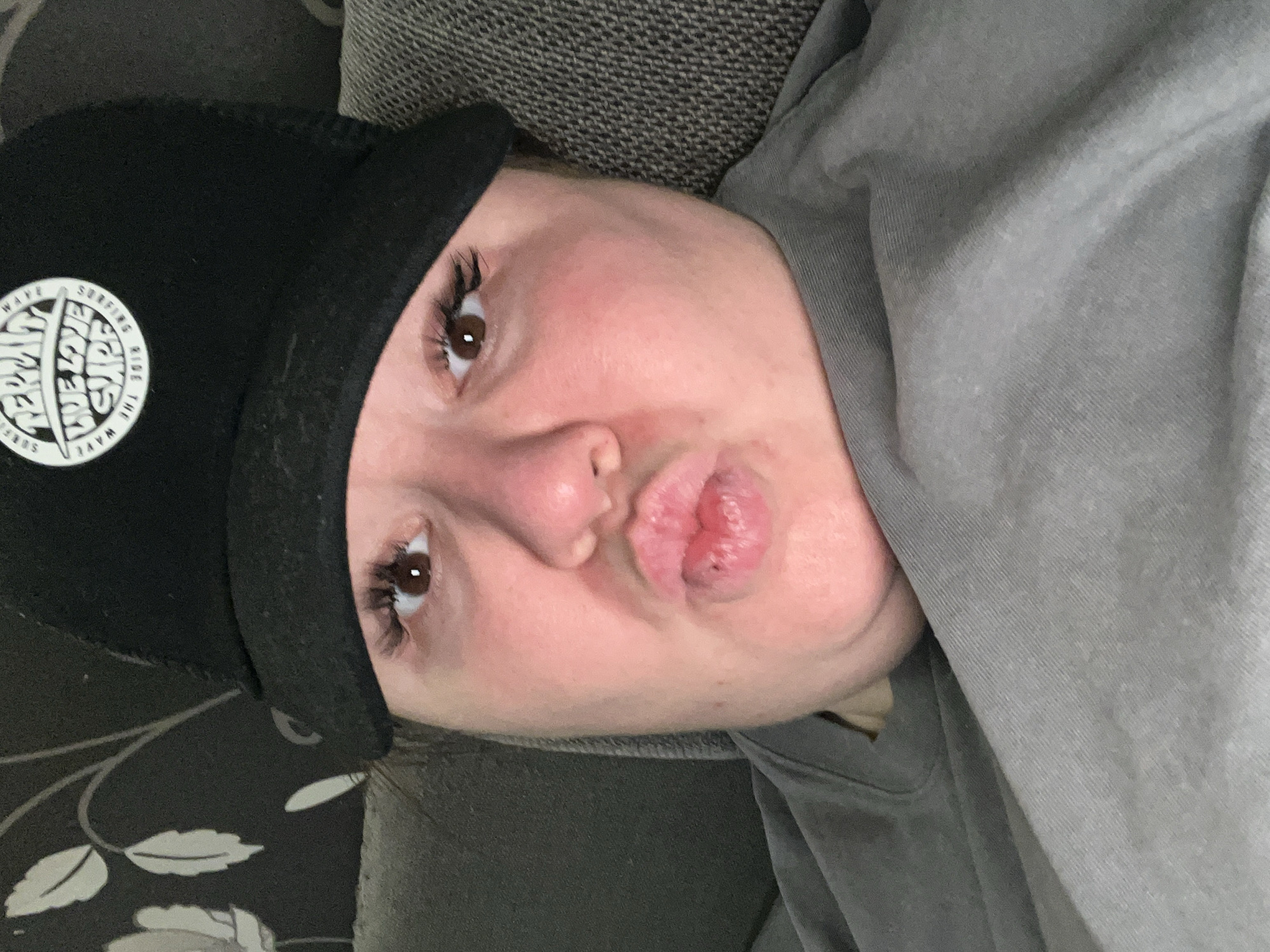An adult individual is depicted in a close-up, head-and-shoulders portrait, appearing to be a selfie. The person, likely female, has a light complexion, brown eyes with prominent eyelashes, and pouted lips. They wear a black baseball cap featuring a circular white logo on the front panel with the legible text "PERM", "TRUE LOVE SURF", "RIDE THE WAVE", and "2018". A grey long-sleeved garment is visible on their upper body. The background on the left consists of a dark fabric or wallpaper with white, stylized leaf or feather patterns, while the right side shows a textured grey fabric, indicative of an indoor setting, possibly a domestic residence. The specific location within the city of Chaikovsky, Russia, cannot be visually determined from the image.