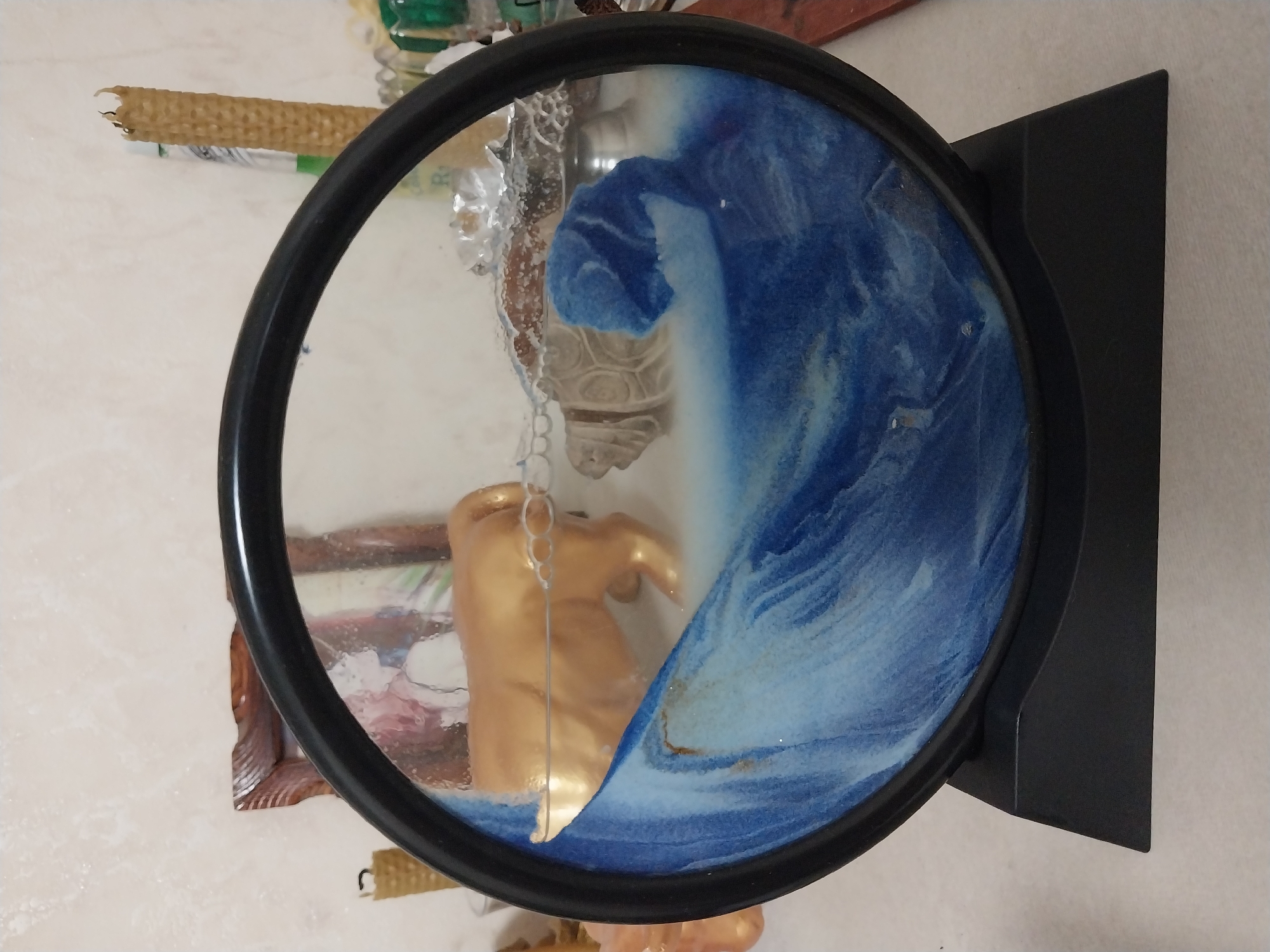 A circular sand art frame sits on a black base. The frame contains a scene with blue and white sand, resembling waves. A golden bull figurine is positioned behind the sand, and a tan turtle figurine is partially submerged in the sand. The frame is placed on a white surface, likely a table, with a textured wall in the background. Candles and other objects are visible in the background, out of focus. The image does not provide any information about the specific location in the city of Dnipro, Ukraine.