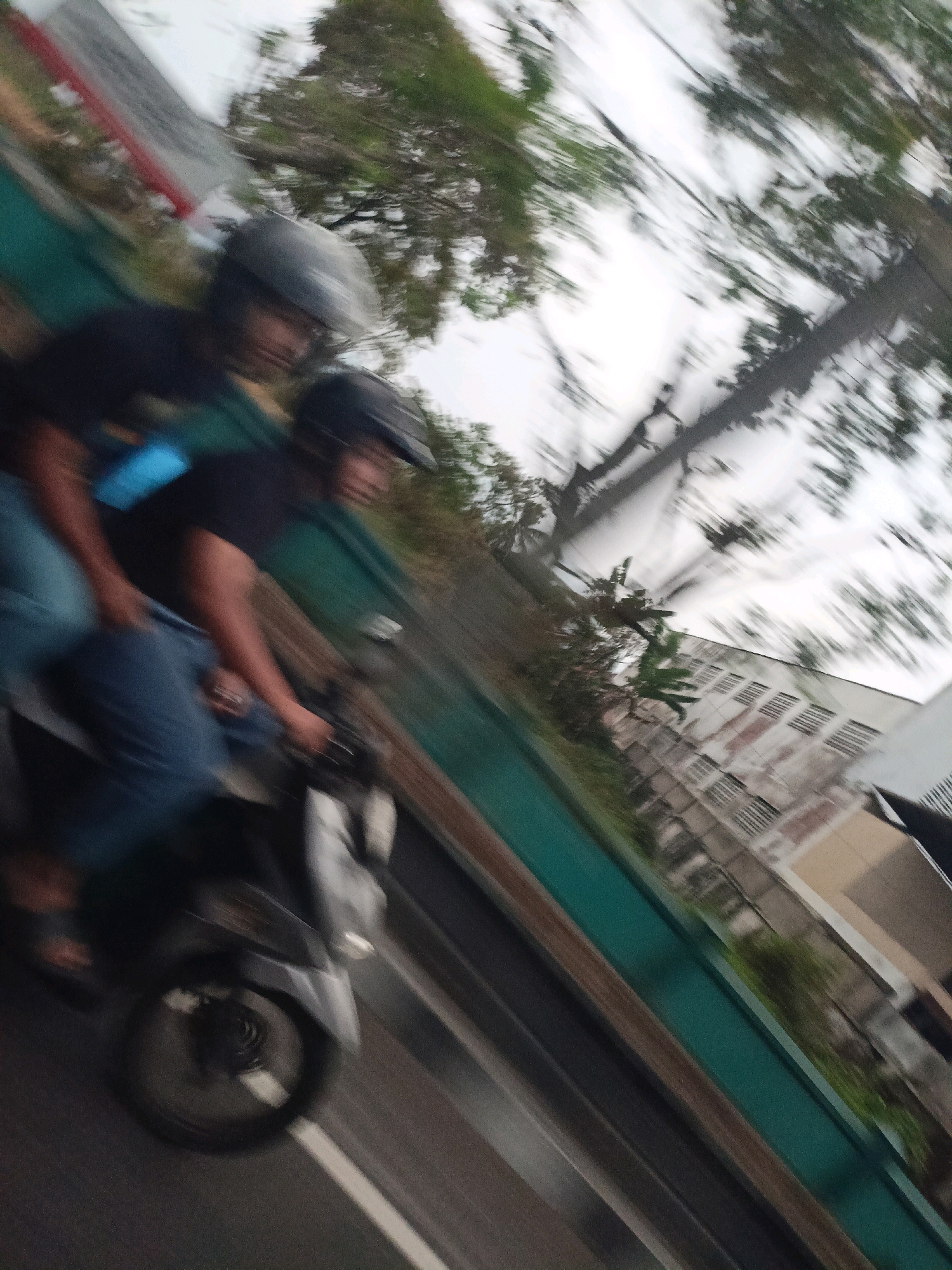 In Payaman, Indonesia, two individuals are riding on a motorcycle, captured in motion blur. The person in front, wearing a black shirt and a gray helmet, appears to be the driver. Behind them, a second individual, also wearing a helmet, is visible. They are dressed in blue jeans. The motorcycle is dark-colored with a silver-colored front fairing. The scene is an outdoor environment, with trees and buildings visible in the background. The angle of the image suggests it was taken from a moving vehicle, possibly another motorcycle or a car, looking towards the right. The blurred motion indicates a fast-moving subject. The lighting appears to be overcast, contributing to the overall soft and blurred appearance of the image.