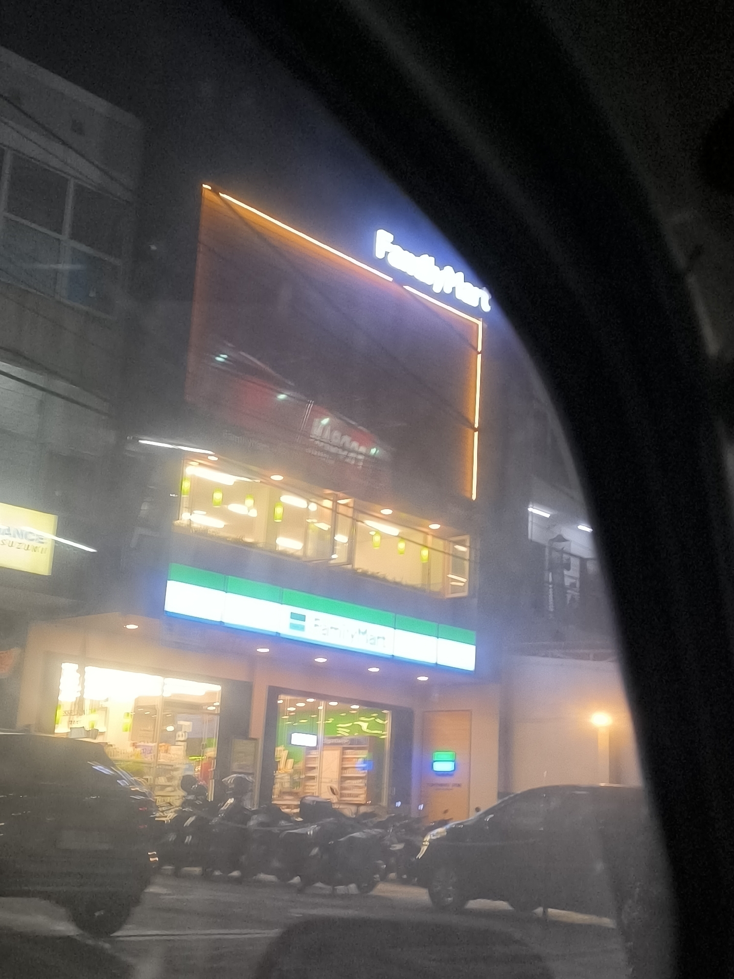 The image depicts a nighttime street scene in Depok, Indonesia, featuring a brightly lit FamilyMart convenience store. The store's exterior signage, including the prominent "FamilyMart" logo in white lettering against a blue background, is clearly visible. The store's windows are illuminated, revealing shelves stocked with products inside. In front of the store, several motorcycles are parked along the sidewalk. A dark-colored car is parked to the left of the motorcycles, partially obscuring the store entrance. The overall environment suggests a commercial area with typical urban nighttime lighting. The image appears to have been taken from inside a vehicle, possibly through a window, as indicated by the dark framing along the edges of the shot.