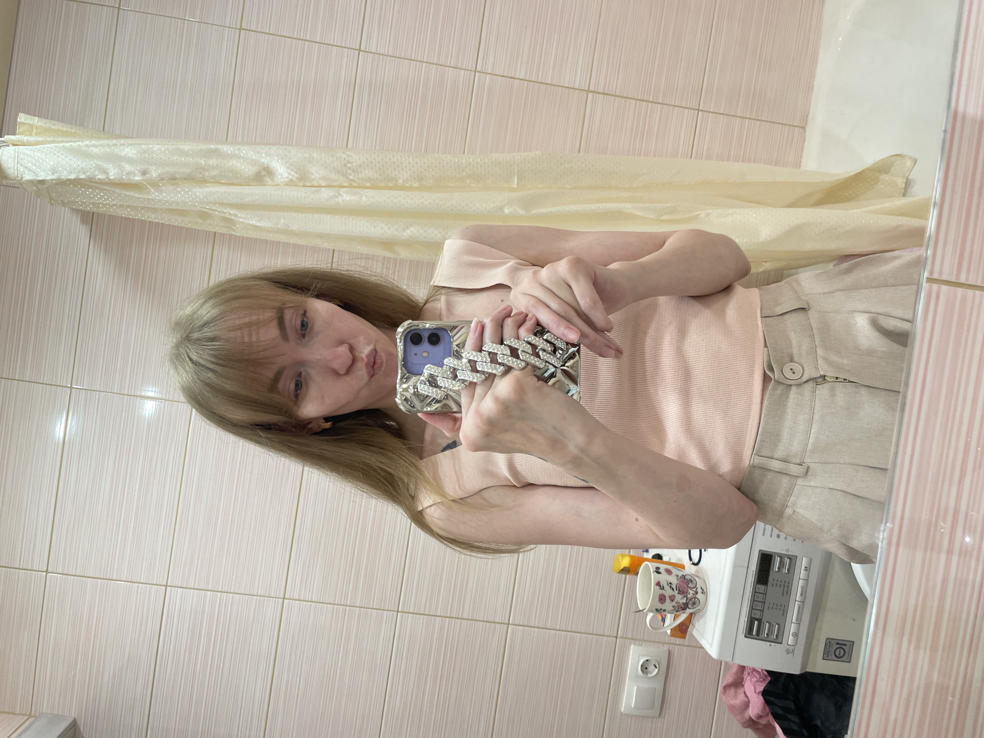 A young woman with long, blonde hair is taking a selfie in a bathroom. She is wearing a light pink tank top and beige shorts. Her phone has a silver chain-link case encrusted with stones. Behind her is a light pink tiled wall, a beige shower curtain, and the edge of a washing machine. There is a teacup on the shelf next to an electrical socket. The scene is likely located in a residential bathroom in Perm, Russia.