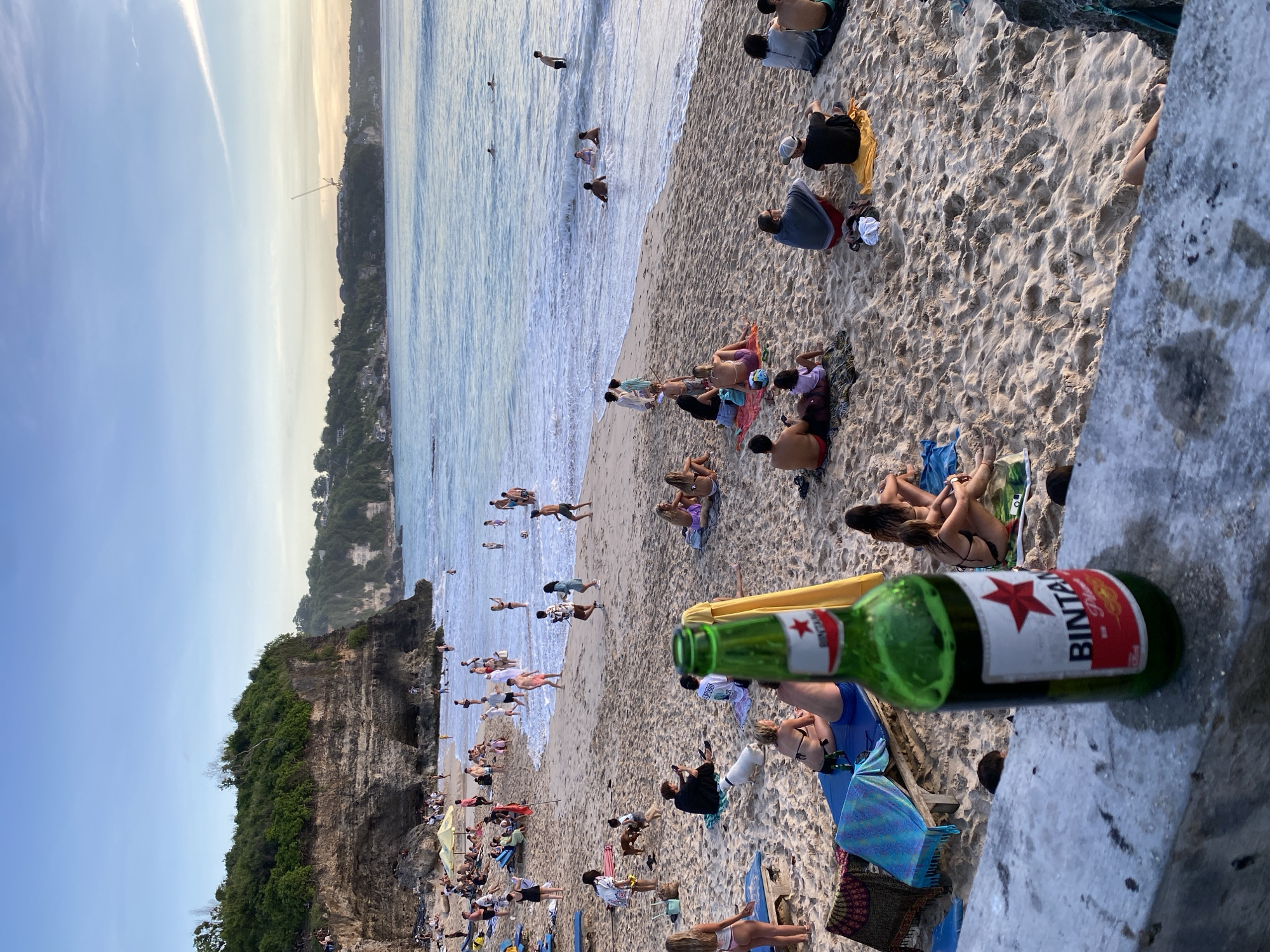 This image captures a vibrant beach scene in Kuta Selatan, Indonesia, during the late afternoon or golden hour, indicated by the warm, soft light illuminating the sand and the sky's gentle transition from clear blue to a lighter, hazy hue near the horizon.
In the immediate foreground, a green glass bottle of "BINTANG PILSENER" beer, with its distinctive red star label, rests on a concrete ledge or wall, slightly out of focus.
The expansive beach is bustling with numerous people. Many individuals are seen relaxing on the light-colored sand, sitting or lying on towels and blankets, socializing or simply enjoying the sun. Closer to the water's edge, several people are wading, swimming, or playing in the ocean's calm, light blue waves. A variety of beach gear, including colorful towels and a partially visible yellow beach umbrella, are scattered among the beachgoers.
To the left side of the frame, a large, dramatic cliff covered in dense green vegetation rises prominently, bordering the beach. The overall setting is that of a popular and lively tropical beach destination, with a relaxed and leisurely atmosphere.