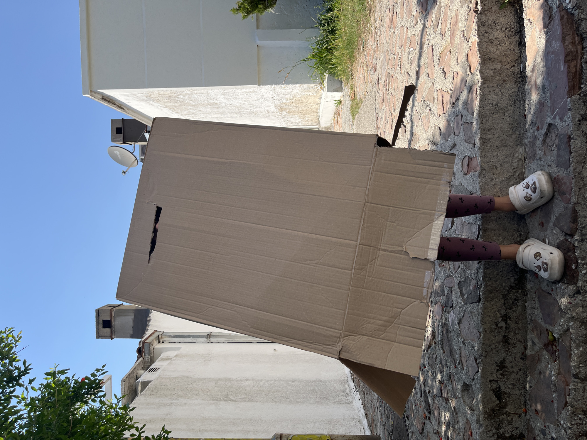 A child wearing patterned leggings and white clogs stands on a stone staircase, partially obscured by a large, angled cardboard box that covers their head and torso. The box has a rectangular opening near the top. The scene is outdoors, with a clear blue sky overhead. To the left, a building with a textured white wall and some green foliage is visible. In the background, there are chimneys and a satellite dish on a rooftop. The ground is paved with stones. The architectural style and presence of a satellite dish suggest an urban or semi-urban environment, possibly in a Mediterranean climate like Budva, Montenegro, given the clear sky and building designs. The lighting indicates a sunny day.