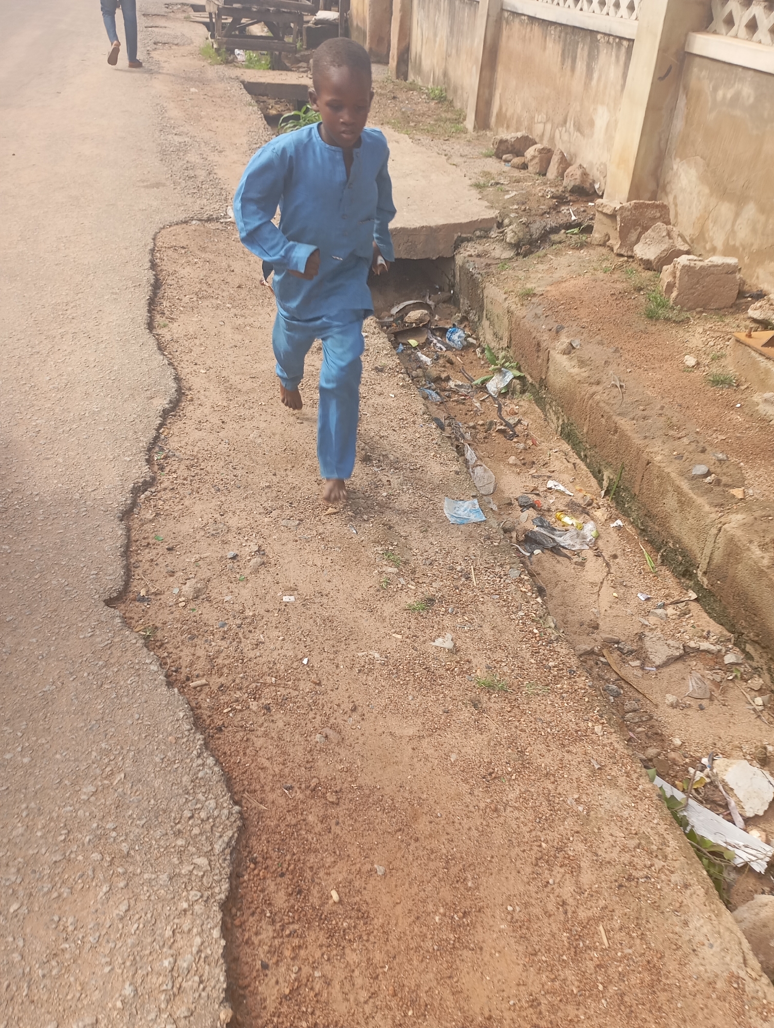 A young boy is running on a dirt path next to a damaged asphalt road in Shendam, Nigeria. The boy is wearing a blue kaftan and appears to be barefoot. To his right, a drainage ditch is filled with debris, including plastic bags, wrappers, and other trash. A concrete wall with decorative elements runs along the far side of the ditch. Further down the road, another person is walking away from the camera, their legs and feet visible. The ground is a mix of dirt, gravel, and some patches of dry grass. The overall scene depicts an outdoor environment with signs of disrepair and litter.