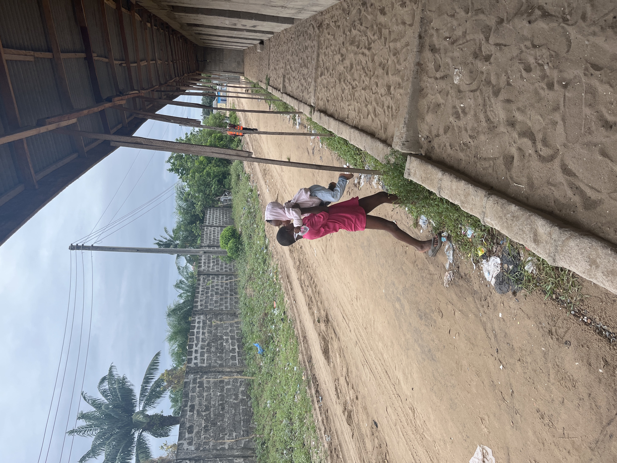 A medium, outdoor shot shows two young people, with a narrow sandy alleyway running alongside a long concrete wall topped by wooden roofing. The picture seems to be taken in Badagry, Nigeria, based on the environment's features.
Two children are visible in the middle of the alley. One child in a pink dress is carrying another child on their back, who is wearing light clothing.
The alleyway is mostly sandy. Litter is present along the bottom edge of the wall. The sandy surface shows tire tracks, footprints, and other disturbances.
The concrete wall is weathered, with some grass and weeds growing along its base. Wooden roofing, supported by posts, overhangs the alleyway along its left side.
Beyond the wall, a brick wall stretches out, with various bushes and trees. A palm tree is visible in the lower-left corner. In the distance, there is a power pole with numerous electric lines. The sky is overcast.