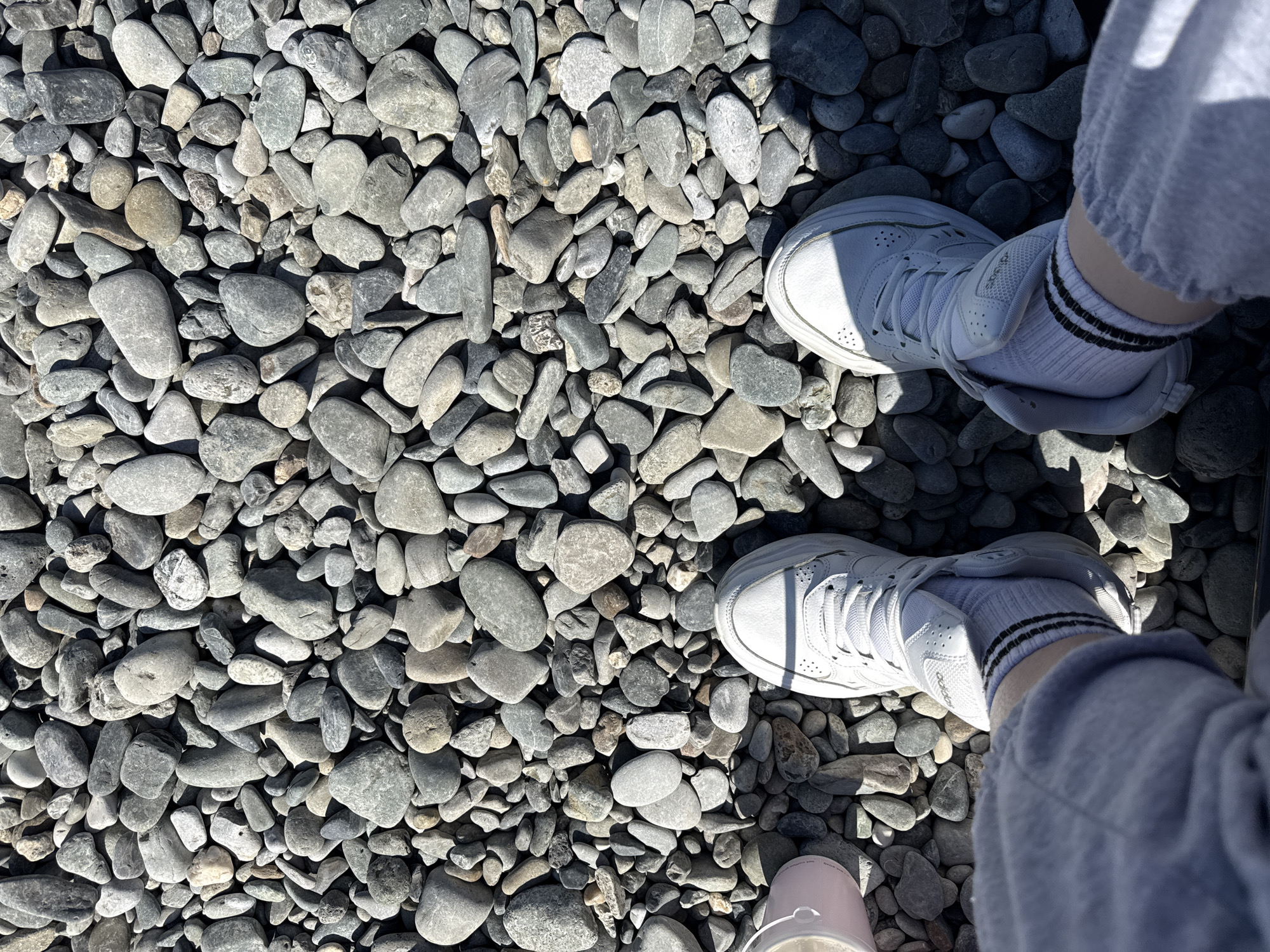 The image presents a high-angle, top-down view of a person's feet standing on a surface covered entirely by numerous smooth, grey and brown pebbles of varying sizes. Bright sunlight illuminates the scene, casting distinct, relatively short shadows from the feet and legs onto the pebbly ground, indicating a sunny daytime setting.
The person is wearing light-colored athletic shoes, likely white or off-white sneakers. Visible above the shoes are white ankle socks featuring three horizontal dark stripes around the cuff. The lower legs are clad in light grey sweatpants or joggers, which appear to have gathered ankles, resting just above the socks.
The brand name "Adidas" is visible in dark lettering on the outer side of the right shoe (which appears on the left side of the frame from the viewer's perspective). A small, light pink object, possibly a cup or another piece of footwear, is partially visible at the very bottom center of the image, with some faint, indecipherable text on its surface. The uniform nature of the pebbles suggests an outdoor environment such as a pebble beach, a riverbed, or a landscaped path.