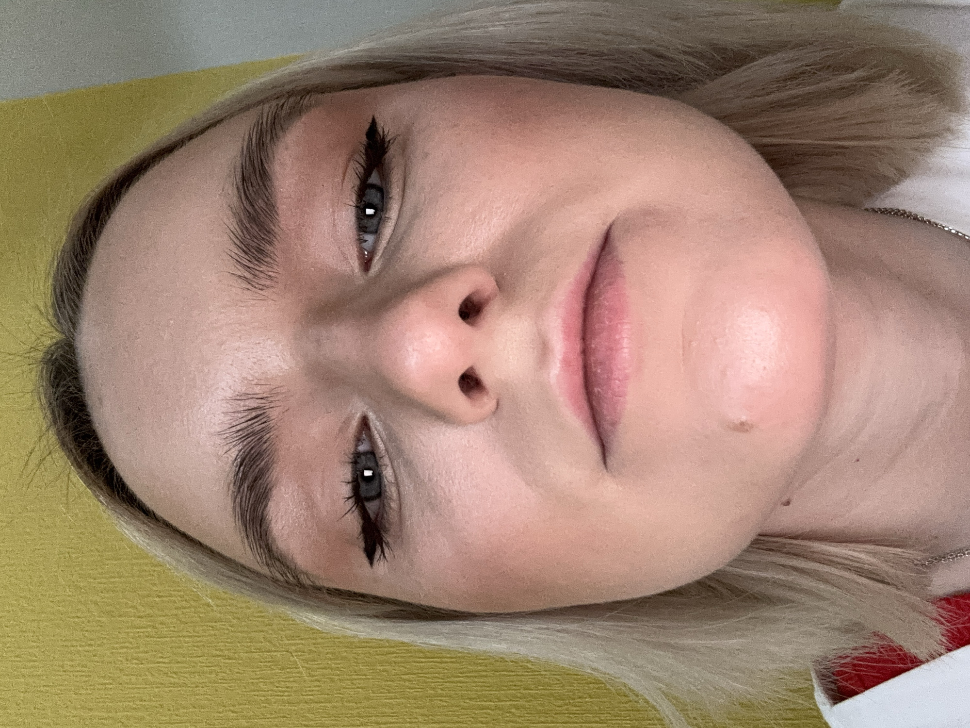 A close-up, angled portrait of a young woman with blonde hair. She has fair skin, dark eyebrows, and wears eyeliner. She is lying down on a surface that appears yellow. A silver chain necklace is visible near her shoulder. The location is not determinable from the image provided.