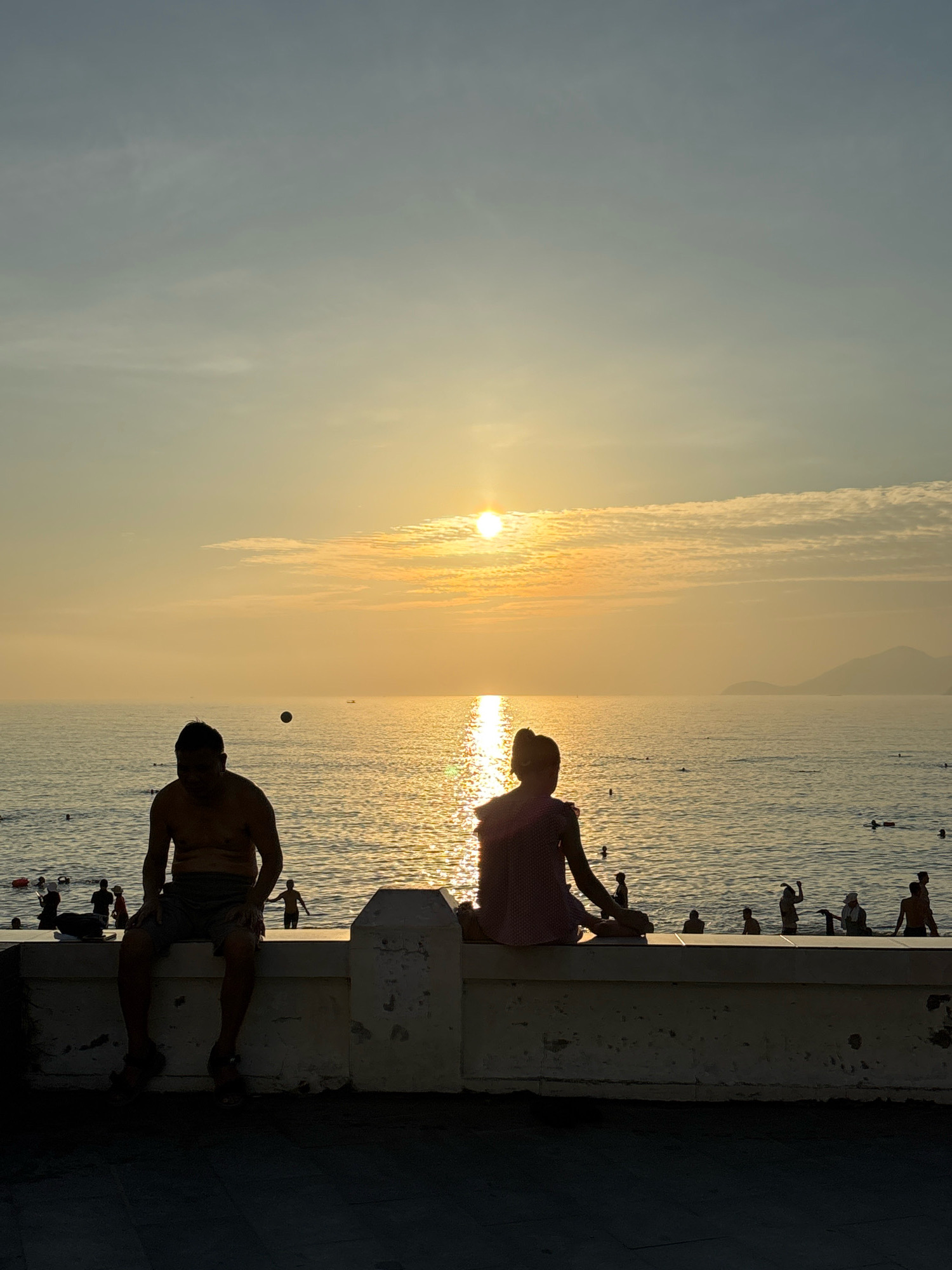 The image depicts a sunset scene at a beach in Nha Trang, Vietnam, likely along the coastline with a promenade. Two silhouetted figures are seated on a low, weathered concrete wall facing the sea. On the left, a man with his back to the camera is seated, appearing to be shirtless and wearing shorts. To his right, a person with their back to the camera, possibly a child or woman, is seated wearing a pink checkered dress. The sun is low on the horizon, casting a warm golden glow across the water, which is dotted with several other silhouetted figures swimming or wading. In the background, a distant shoreline with mountains is visible, and a few small boats are on the water. The overall mood is serene and tranquil, capturing a typical evening at a Vietnamese beach.
