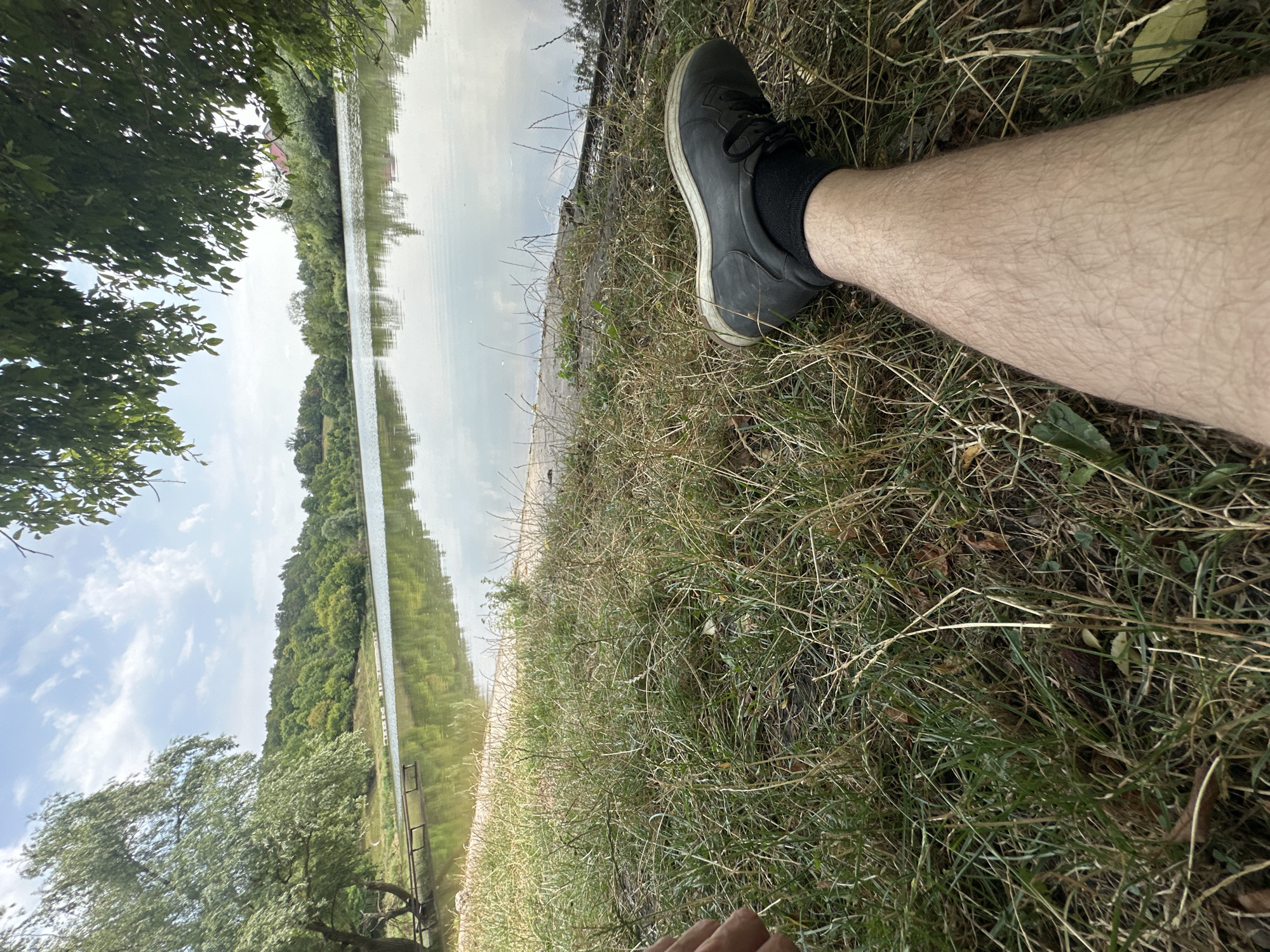 A low-angle, close-up shot captures a person's hairy leg and foot resting in overgrown grass near a tranquil lake. The leg is clad in a black sock and a black shoe with white soles. The lake, reflecting the sky and surrounding lush green trees, stretches into the distance. A wooden jetty is visible on the left bank. The sky is partly cloudy with a bright, hazy sun. The scene is set in an outdoor, natural environment, likely a park or lakeside area, in Makiv, Ukraine.