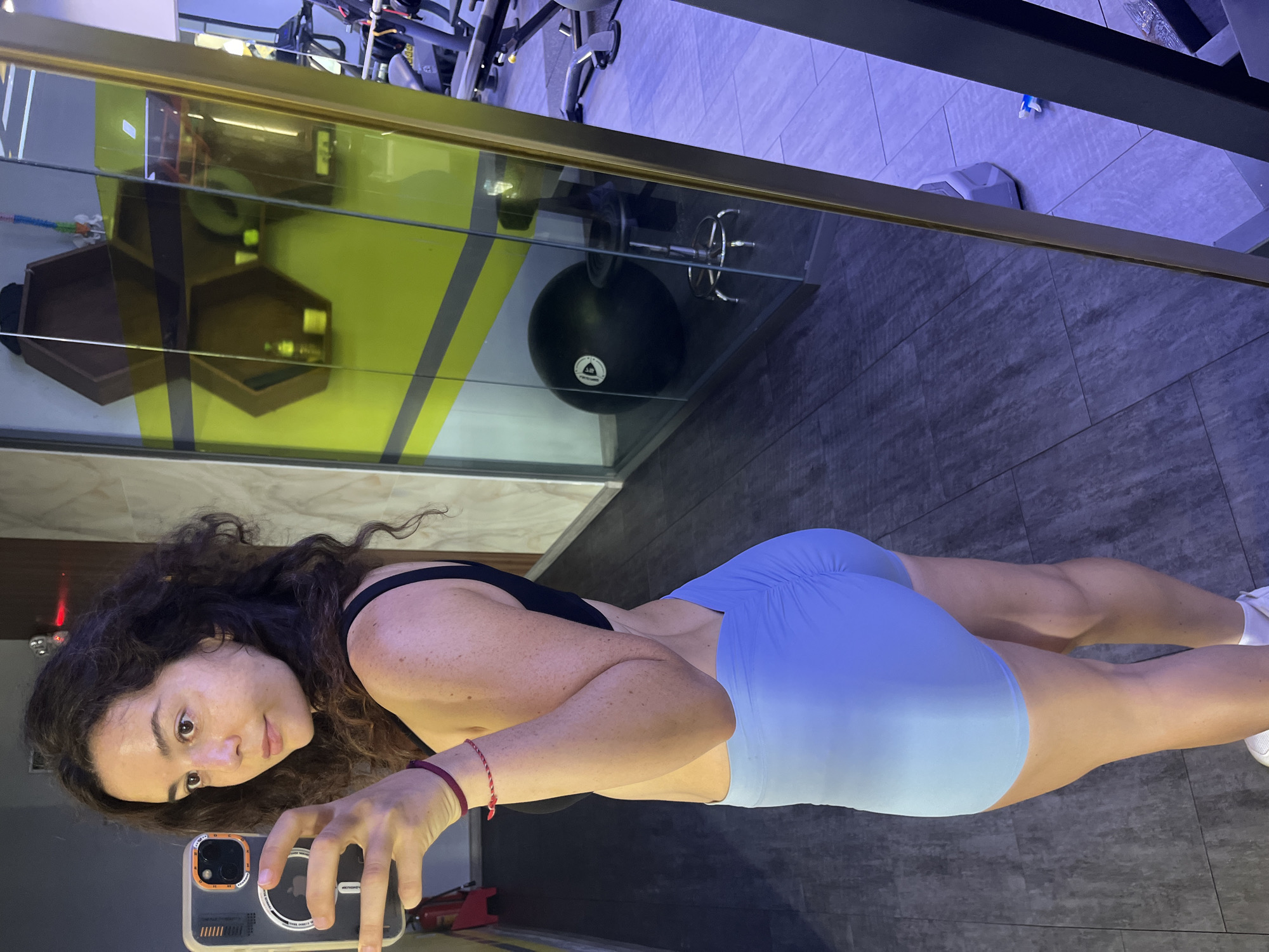 A young woman with curly dark hair stands in a gym, taking a selfie in a mirror. She is wearing a black sports bra and light blue shorts. The gym has a modern design with gray tiled floors and yellow and gray accent walls. Reflecting in the mirror are various gym equipment, including weight machines, a large black exercise ball, and a small stool. The lighting in the gym appears to be a mix of natural and artificial light, creating some shadows. The overall scene suggests a workout session or a moment of personal reflection at a fitness facility. The location specified, Phu Thuy Ward, Vietnam, provides a geographical context for this scene.