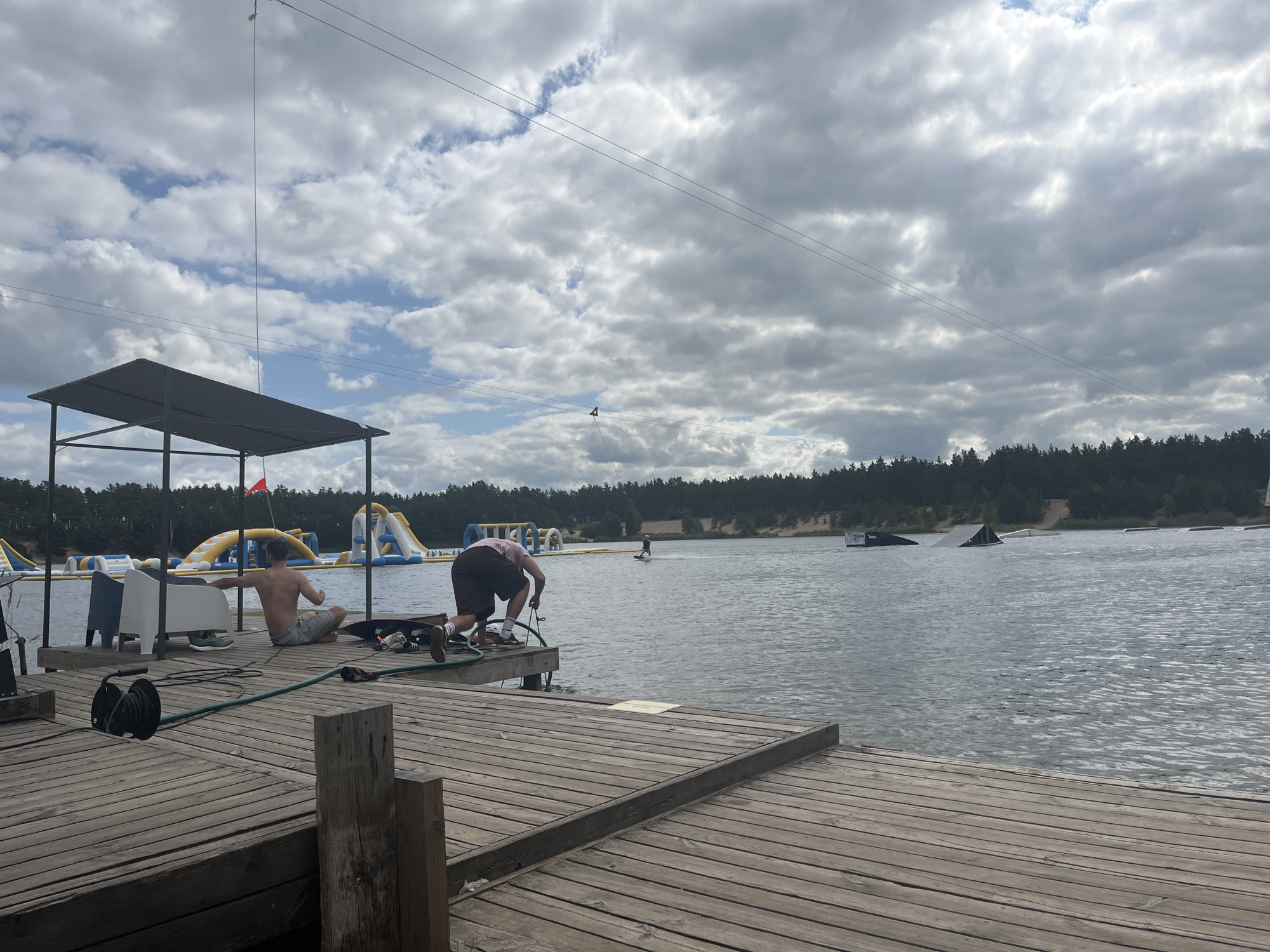 The image depicts a wakeboarding park on a cloudy day in Männiku, Estonia. In the foreground, a wooden pier extends into the water, with two individuals on it. One person, shirtless and wearing light-colored shorts, is seated with their back to the camera, facing the lake. Another person, wearing dark shorts and a pink shirt, is bending over and appears to be preparing for wakeboarding. A wakeboard and a hose reel are visible on the pier. In the middle ground, the lake features several inflatable water obstacles and ramps. Further in the background, a dense forest lines the shore. Overhead, power lines are strung across the frame against a sky filled with dramatic clouds.