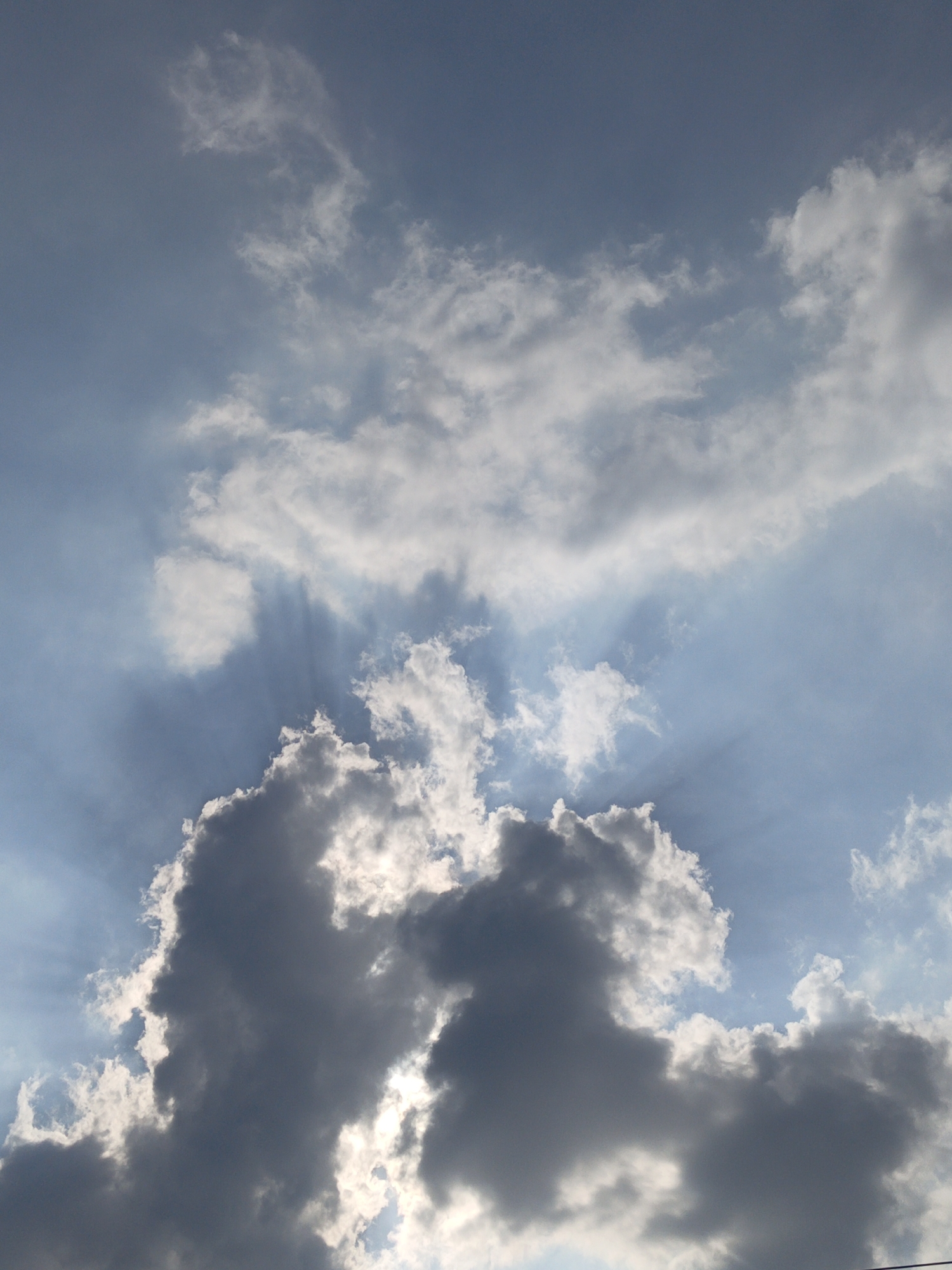 The image depicts a sky filled with a dynamic cloudscape. A large, dark cumulonimbus cloud dominates the lower portion of the frame, with its textured, shadowed mass contrasting sharply against the bright, sunlit sky. Shafts of light, known as crepuscular rays, pierce through gaps in the clouds, creating dramatic beams that extend upwards. Lighter, more dispersed clouds are visible in the upper areas of the sky, some appearing wispy and translucent, while others are more substantial. The overall impression is one of a partly cloudy day with the sun breaking through. There are no people, structures, or recognizable landmarks visible in this image, making it impossible to determine the specific location within Gedung Nyapah, Indonesia, or any particular activity. The scene is purely atmospheric.