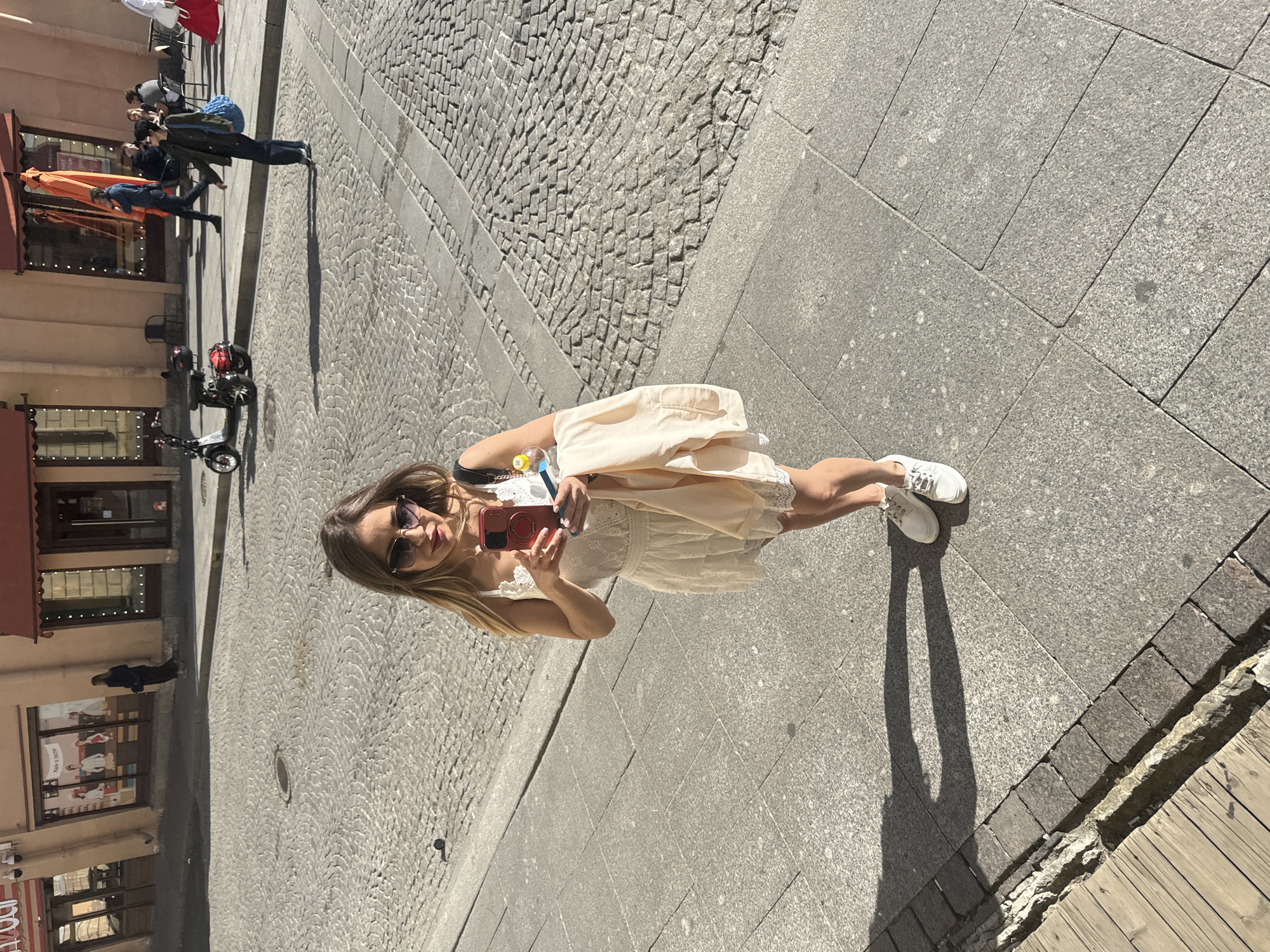 A blonde woman wearing a white lace dress, white sneakers, and sunglasses is standing on a paved city street and taking a picture with her phone. She is holding a peach-colored jacket over her left arm. The street is paved with cobblestones and large granite slabs. In the background, there are buildings with storefronts, and several people are walking in the distance. A scooter is parked on the street. The sun is casting strong shadows. The scene appears to be in an urban area of Saint Petersburg, Russia, possibly near a historic district or a shopping area given the architecture of the buildings and the style of the paving.