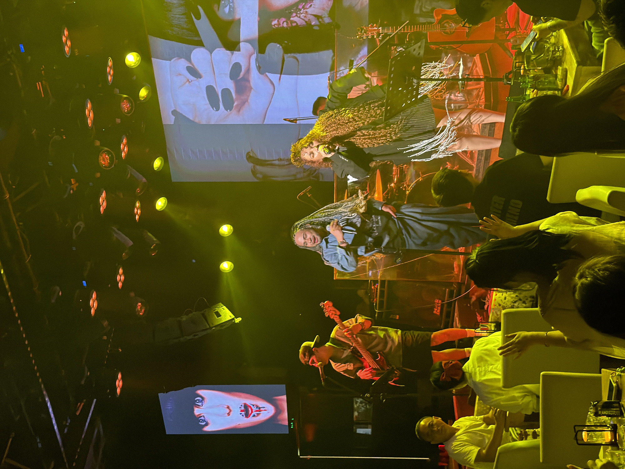 In a dimly lit venue, possibly in Jiangbeicheng Street, China, a musical performance is underway. On stage, a woman with long, curly auburn hair is singing into a microphone, wearing a black and white striped outfit adorned with fringe. Beside her, a person with long dreadlocks and wearing a blue denim-like outfit is also holding a microphone. A guitarist in a light-colored shirt and shorts, wearing a cap, plays a red electric guitar. Behind the performers, a large screen displays images, one featuring close-ups of hands with long black nails and another abstract digital art. The stage is illuminated by warm yellow spotlights and overhead lighting fixtures. In the foreground, audience members are seated at tables, some looking towards the stage, others engaged with their phones. The overall atmosphere suggests a lively indoor concert or club setting.