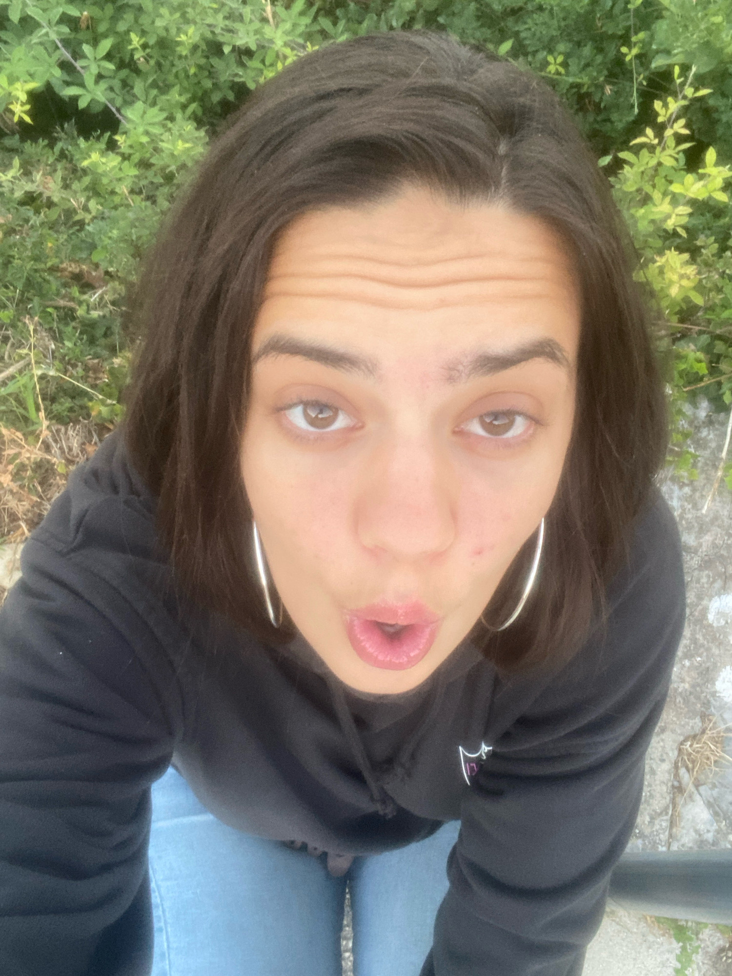 A young woman is captured from a high-angle perspective in a selfie, with her face turned towards the camera. She has dark brown hair, styled loosely around her face, and is wearing large silver hoop earrings. Her expression is one of surprise or playful astonishment, with her mouth open and eyebrows raised. She is dressed in a black hooded sweatshirt and light blue jeans.
The background is blurred and consists of lush green foliage, suggesting an outdoor setting. The ground beneath her appears to be a mix of rough, greyish-white stone and dry grass. There are no other people or discernible objects in the immediate vicinity.
This image was taken on the outskirts of the city of Kučiće, Croatia, likely in a natural or semi-natural environment. The lighting suggests it is daytime, with soft, diffused light. The overall impression is casual and spontaneous.