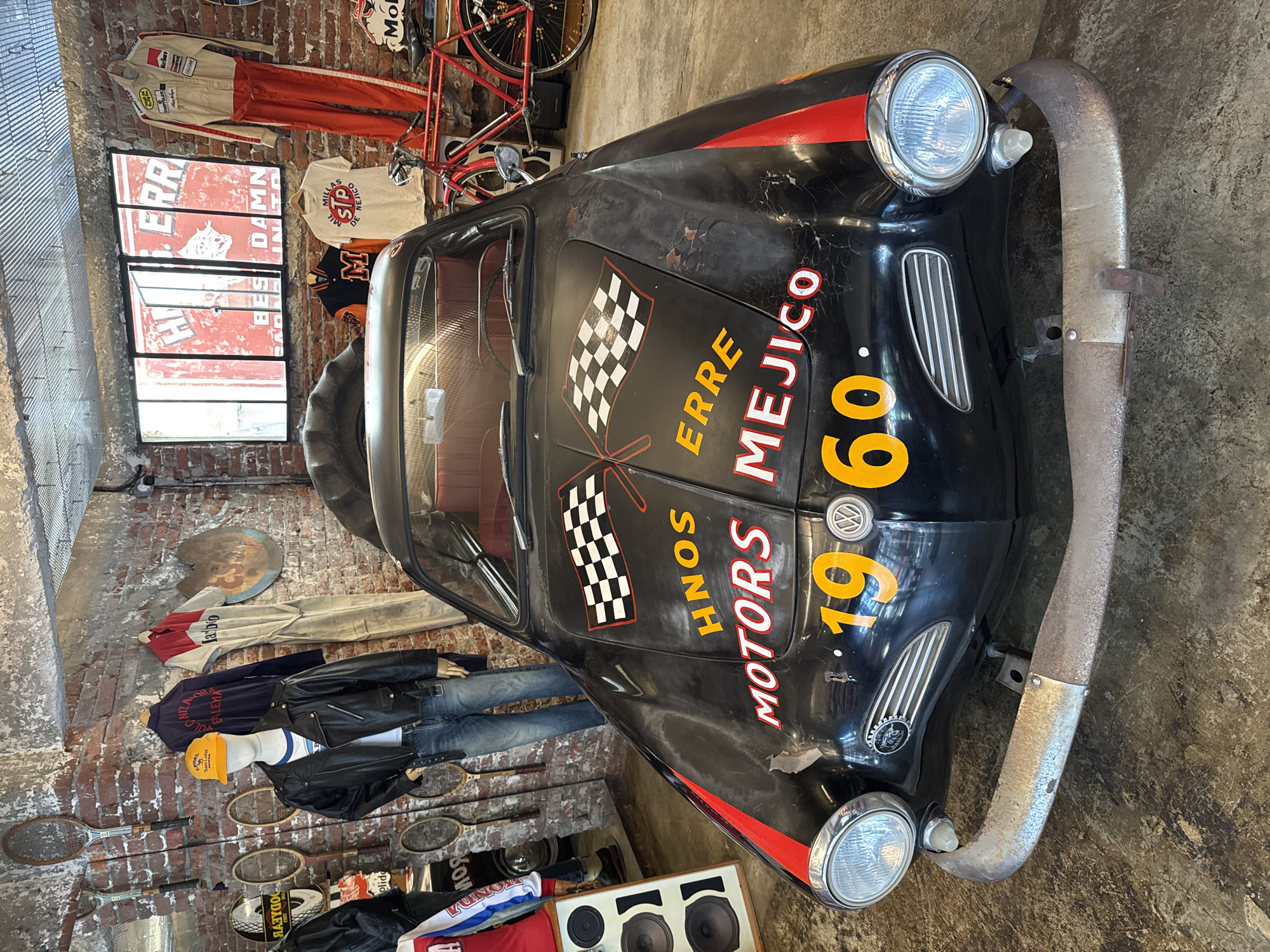 The image shows a black vintage Volkswagen Karmann Ghia race car, likely in Mexico City, Mexico, given the inscription "MEJICO" on its hood. The car features racing-style decals including checkered flags, the text "HNOS ERRE MOTORS MEJICO," and the number "1960." The backdrop includes a brick wall decorated with various items: racing suits, clothing on mannequins (including a leather jacket, jeans, and t-shirts), tennis rackets, speakers, and other racing-related memorabilia. A red bicycle is visible at the top of the image. The lighting is a mix of natural light from a window and artificial light, adding to the overall vintage feel of the scene.
