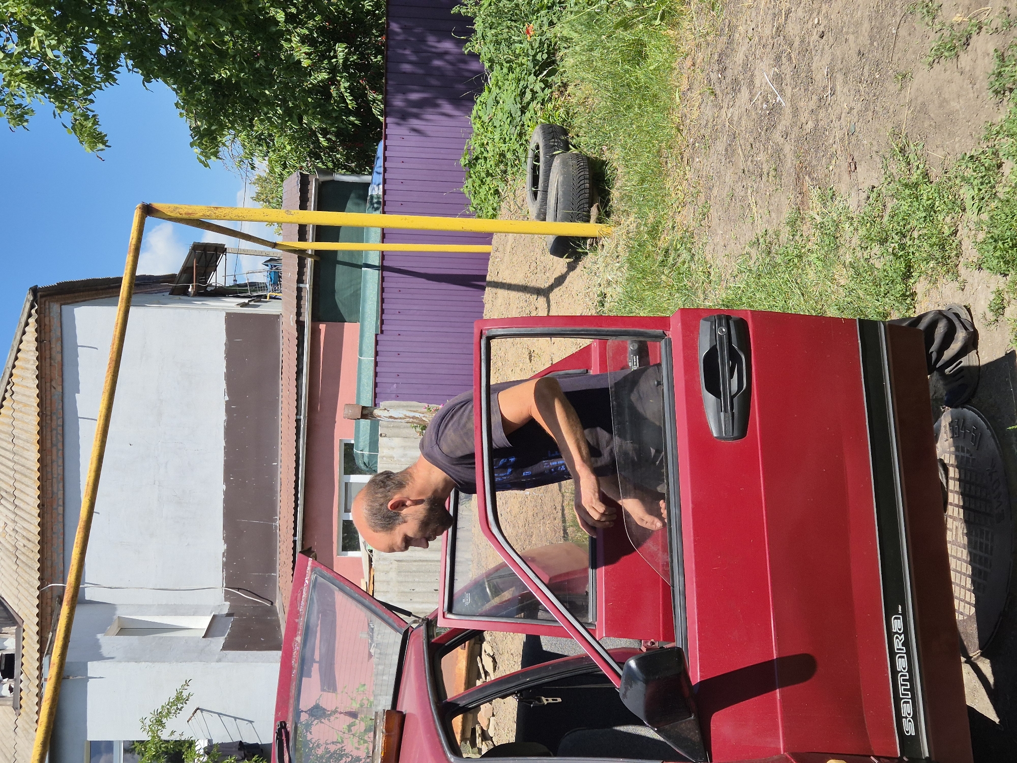 A bald man with a beard, wearing a dark t-shirt, is standing next to an open red car, with his body partially inside the vehicle. The car has its driver's side door and rear passenger door open. The background shows a building with a corrugated roof, a purple metal fence, green foliage, and a yellow metal structure. The ground is a mix of dirt, grass, and scattered rocks. Two tires are visible near the fence. The scene appears to be in an outdoor, possibly rural, setting within the city of Sozonivka, Ukraine.