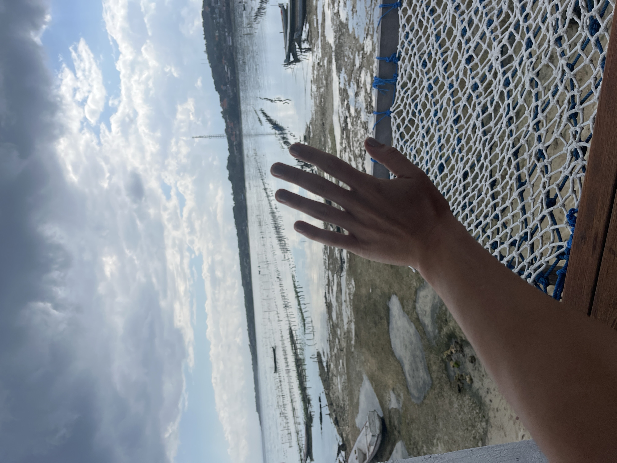 A hand is held up in the foreground, obscuring part of the view. Beyond the hand is a body of water with a shoreline of sand and rocks. Numerous poles are planted in the water, forming rows that extend towards the horizon. Two small boats are visible in the water, one closer to the foreground and another further away. The background shows a distant coastline with vegetation and buildings, and a telecommunications tower. The sky above is cloudy with patches of blue. The scene appears to be a coastal area, possibly a farm or cultivation site for seaweed or shellfish, in Nusa Penida, Indonesia.
