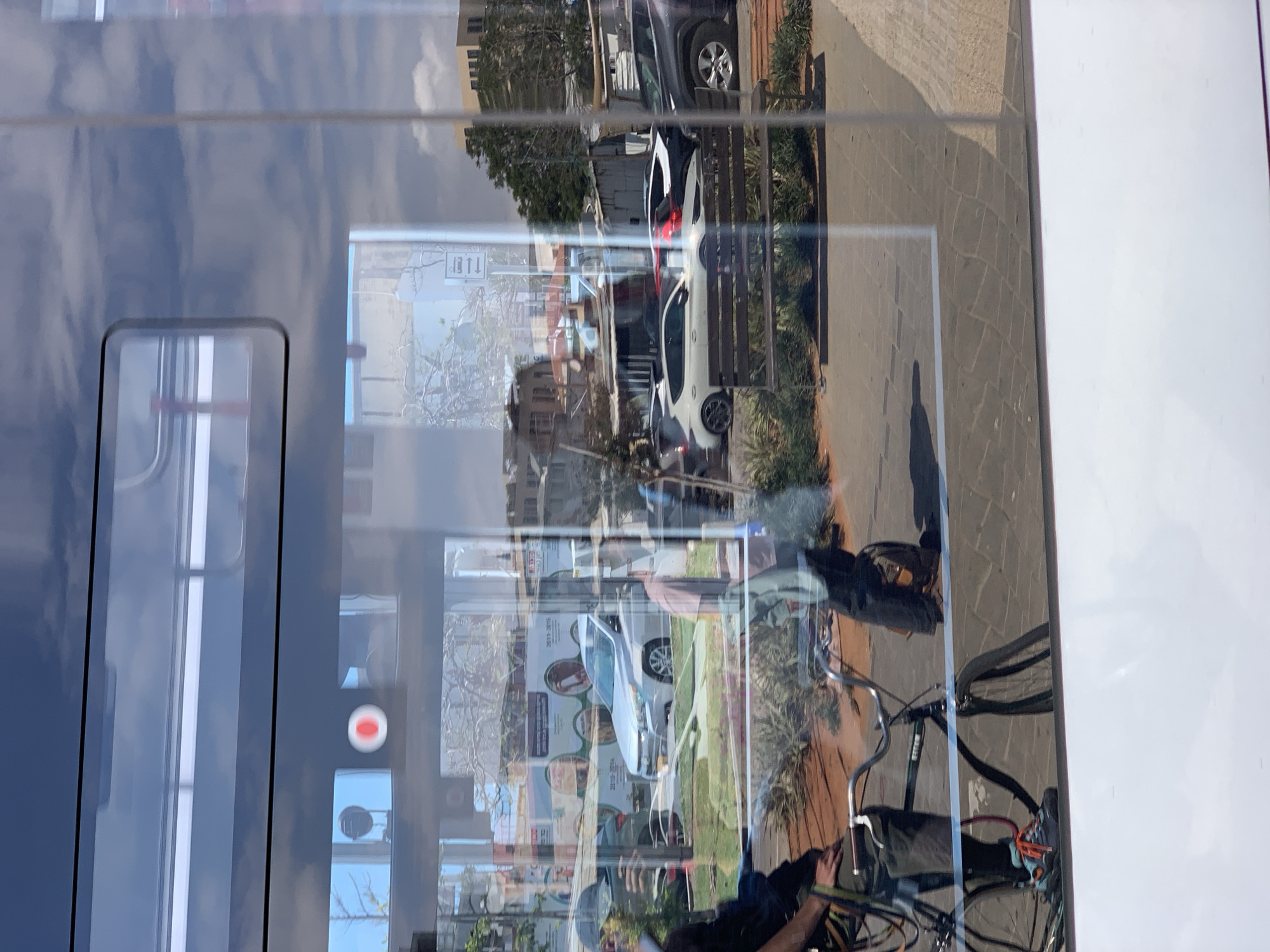 The image captures a scene through a window, likely of a train or bus. Reflections on the window show an urban environment, including streets, cars (a white car, a gray Jeep), buildings, and a pedestrian area with paving stones. In the foreground, a person on a bicycle is partially visible. The reflections suggest that the location is in Tel Aviv, Israel. There is no direct interaction among the subjects, as the person on the bicycle is outside the vehicle reflecting the city view.