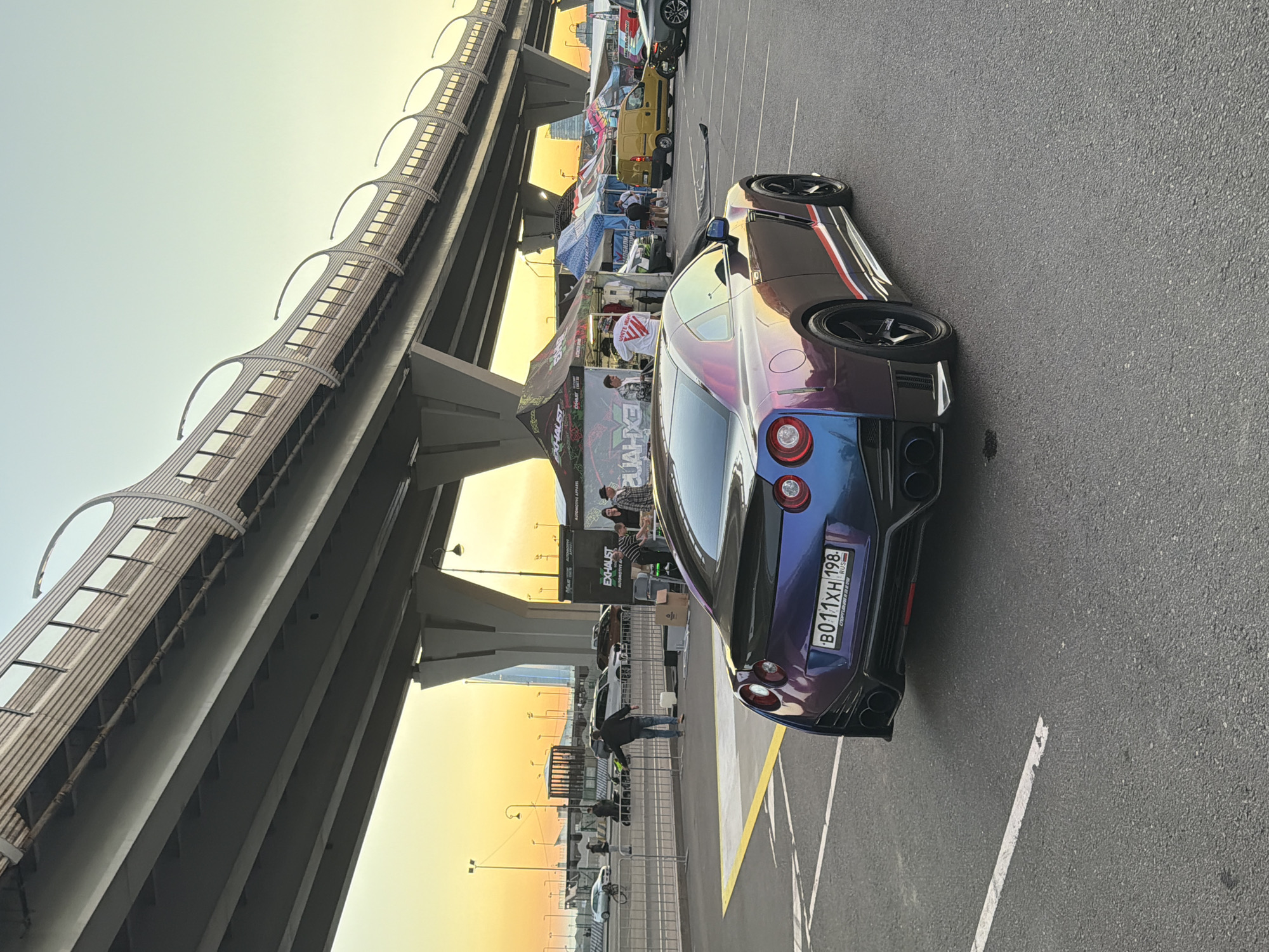 An eye-level shot captures a customized Nissan GT-R parked on an asphalt surface, likely a parking lot or event space. The car, with a distinctive iridescent paint job shifting between purple, blue, and pink, faces away from the camera, angled slightly to the right. Its rear features prominent exhaust tips and a spoiler. The car's license plate, "B011XH 198 RUS," is visible.
Behind the car, a row of tents, possibly for an automotive event, are set up under an elevated highway structure. The highway's concrete supports and ribbed underside are prominent in the background. Several individuals are milling around the tents and a yellow van parked nearby. A metal barrier runs along the edge of the parking area, separating it from a paved walkway. Further in the background, a white car is parked, and a bicycle is leaning against a fence. The scene is illuminated by the warm glow of what appears to be sunset or sunrise, casting a soft light on the environment. The sky transitions from a pale yellow at the horizon to a light blue.