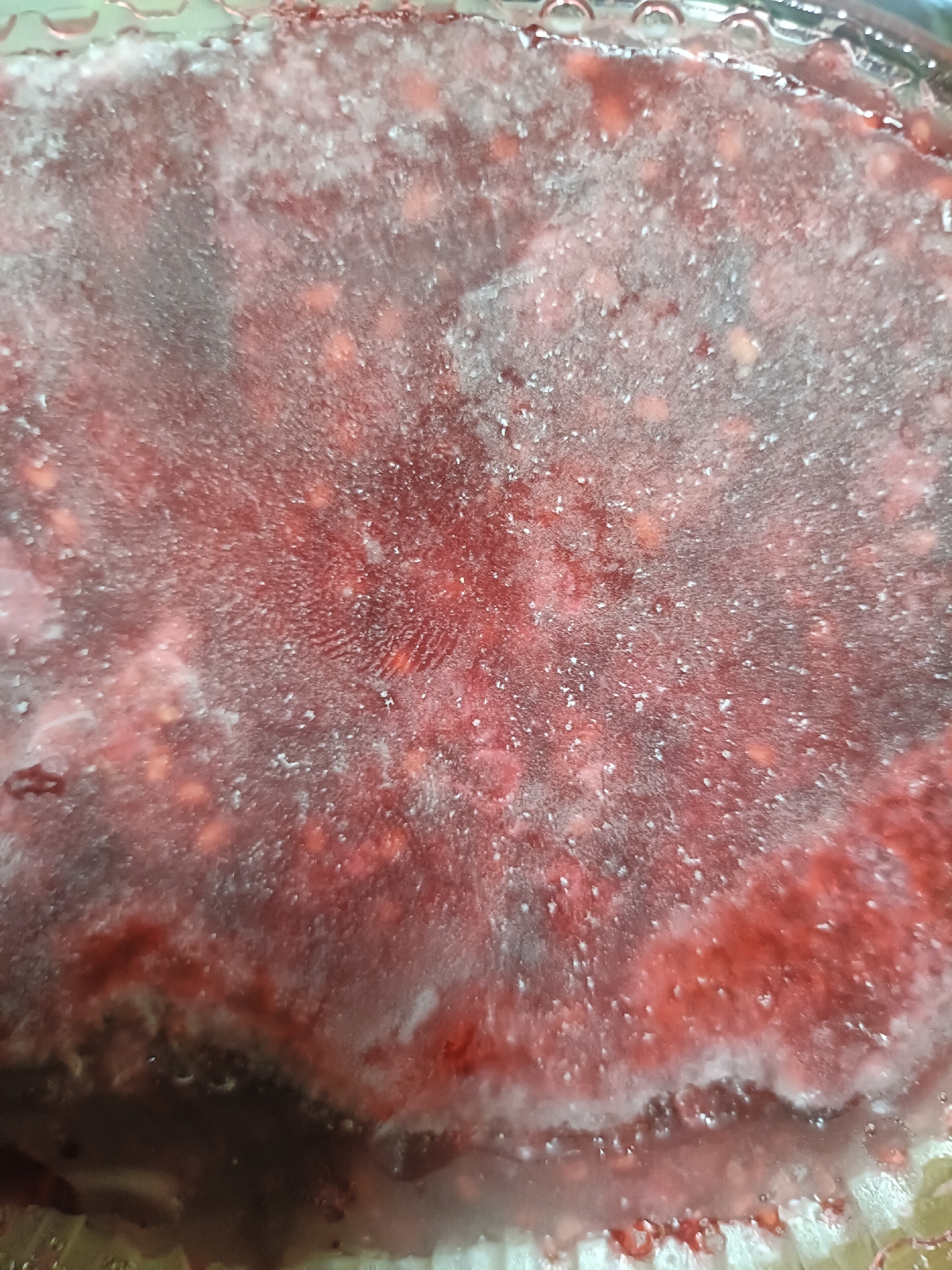 The image shows a close-up, overhead view of a frozen solid substance with a reddish-pink hue, likely berries, contained within a clear glass dish. The surface of the frozen substance is covered with a layer of frost and ice crystals, creating a slightly opaque appearance. Some small, lighter-colored particles are visible within the frozen mass, which could be seeds or other fruit components. The texture appears somewhat granular due to the ice. The edges of the glass dish are visible, showing some condensation or frozen moisture along the rim. The lighting creates some glare and highlights on the icy surface.