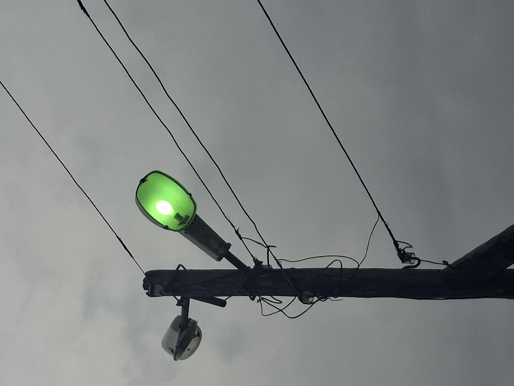 A green street light is mounted on a utility pole under a gray, overcast sky. Electrical wires run diagonally across the top of the frame. A smaller, unlit street light is also attached to the pole. The exact location within Khlevnoye, Russia, cannot be determined from the image. No people or actions are visible.