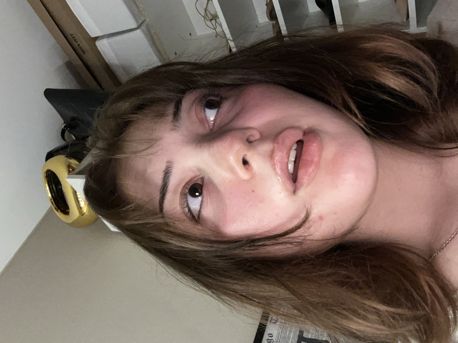 A person with brown hair and light skin looks up. The person's lips are slightly parted, showing their teeth. Their eyes are brown and look towards the upper left. A gold object sits near their head. Boxes and wires are in the background.
